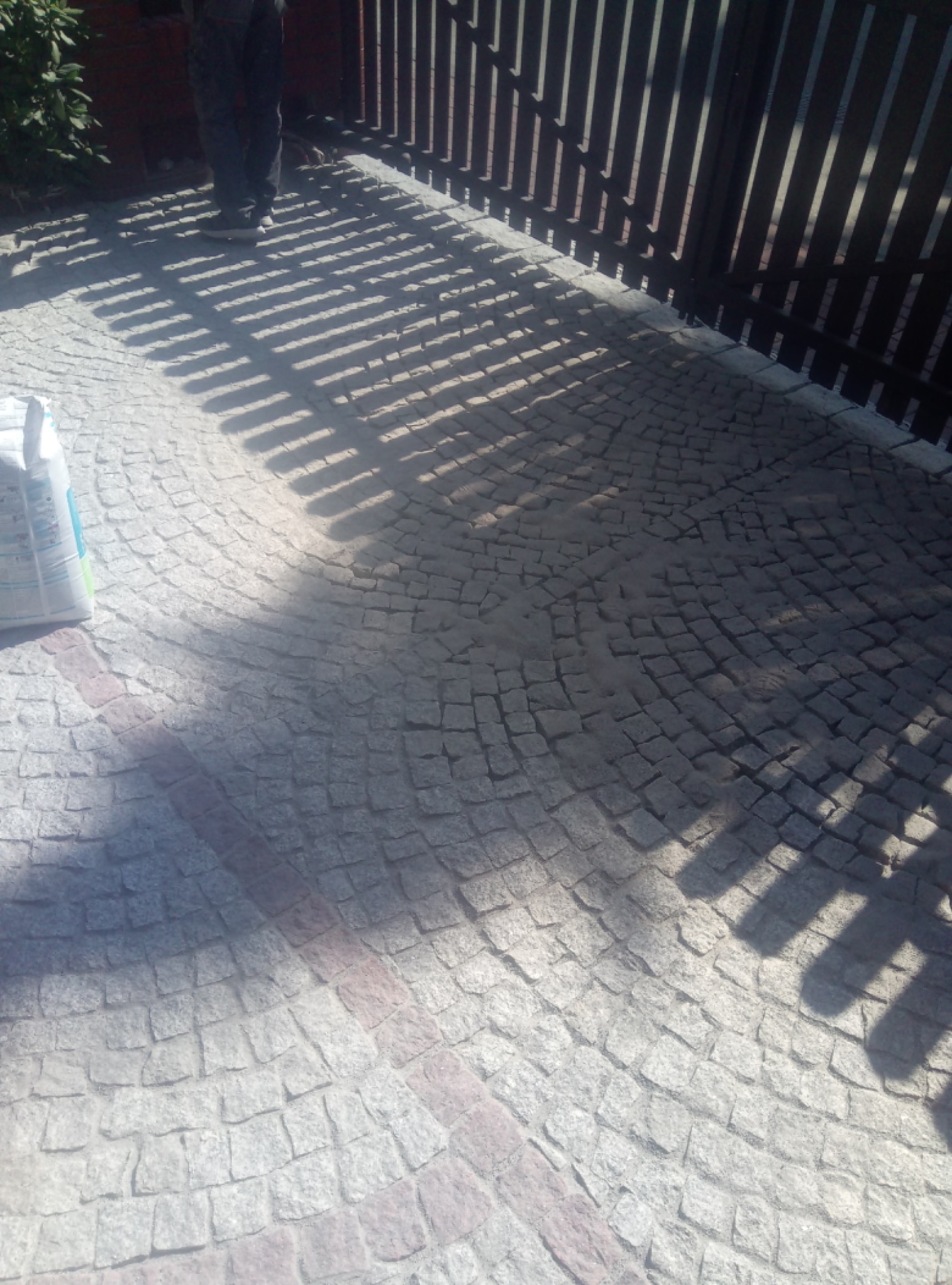 Ułożona kostka brukowa w kształcie wachlarza z widocznymi cieniami płotu i częścią sylwetki osoby oraz workiem z fugą.