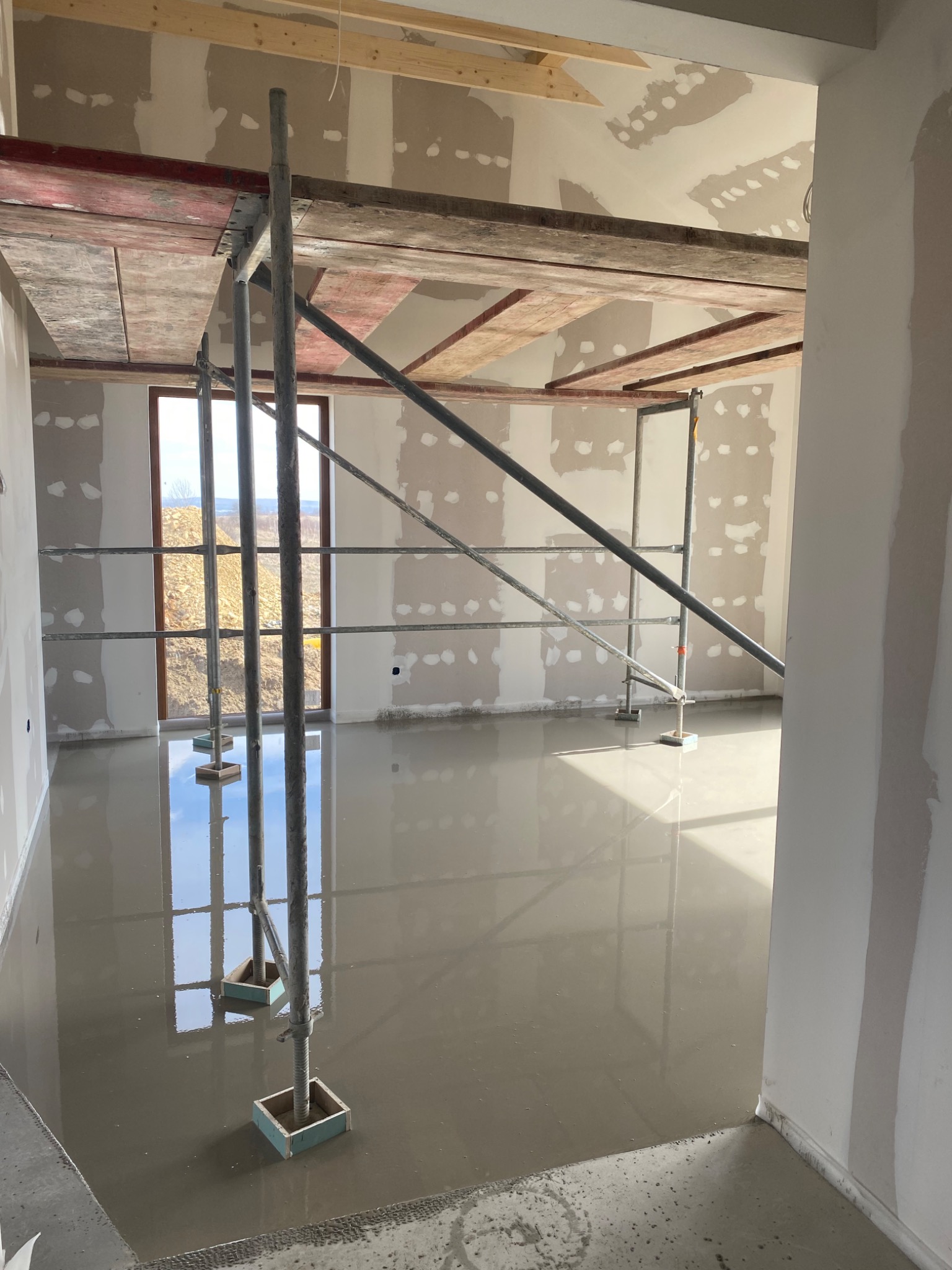 Wnętrze w trakcie wylewki samopoziomującej; widoczne rusztowanie, okno z widokiem na zewnątrz i ściany z widocznymi łatami gipsowymi.