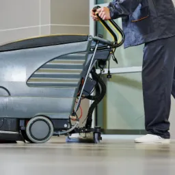 Osoba w uniformie obsługuje profesjonalną maszynę do czyszczenia podłóg w jasnym, nowoczesnym biurze z szarymi płytkami podłogowymi.