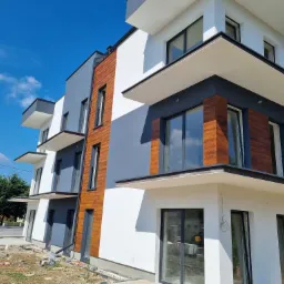 Nowoczesny budynek wielorodzinny z elewacją w kolorach białym, szarym i drewnopodobnym, z widocznymi balkonami i dużymi oknami, sfotografowany w słoneczny dzień na tle błękitnego nieba,...