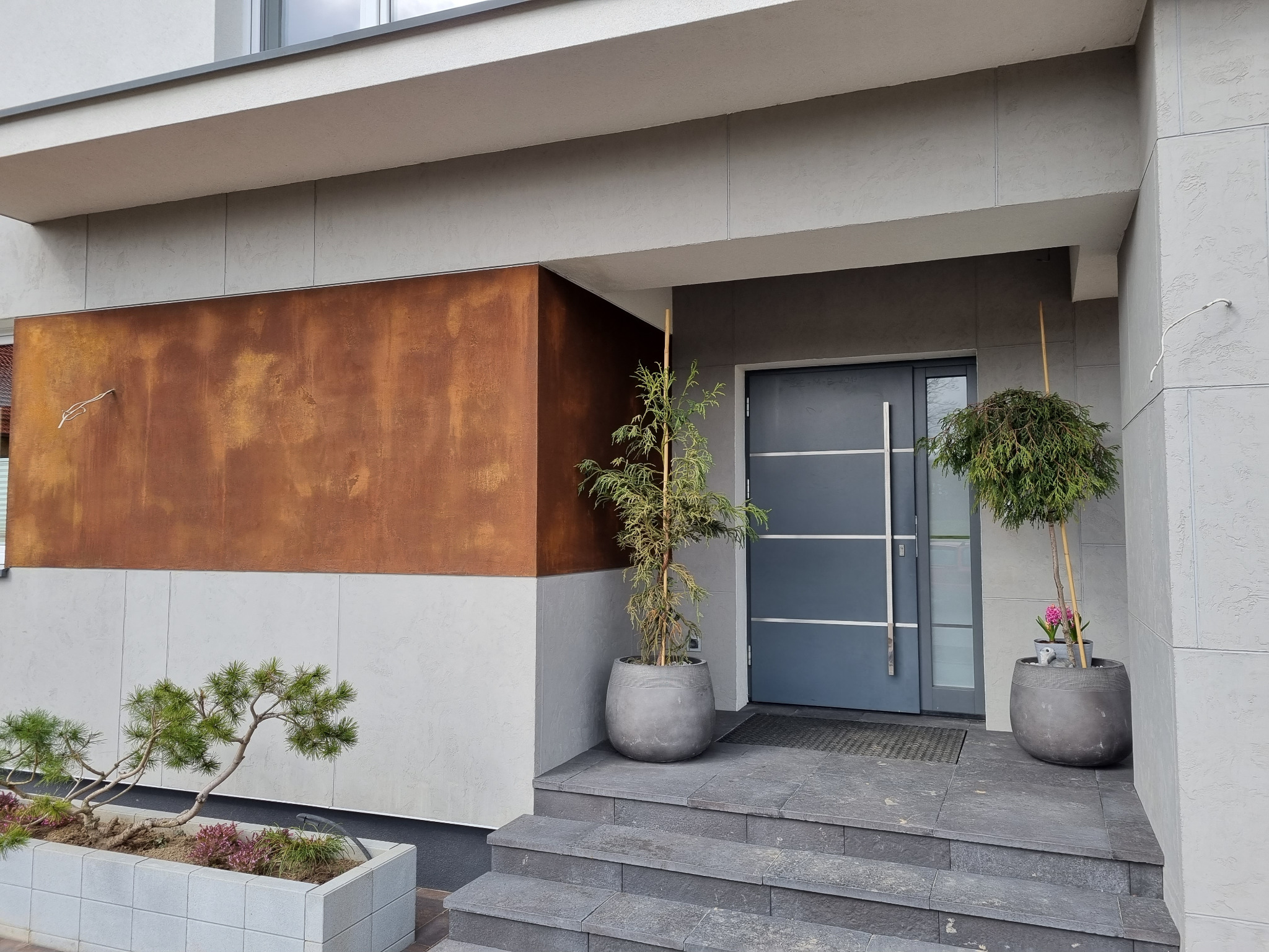 Nowoczesna elewacja domu z elementami dekoracyjnymi: rustykalna płyta oraz szara struktura tynku, wejście z roślinami w donicach.