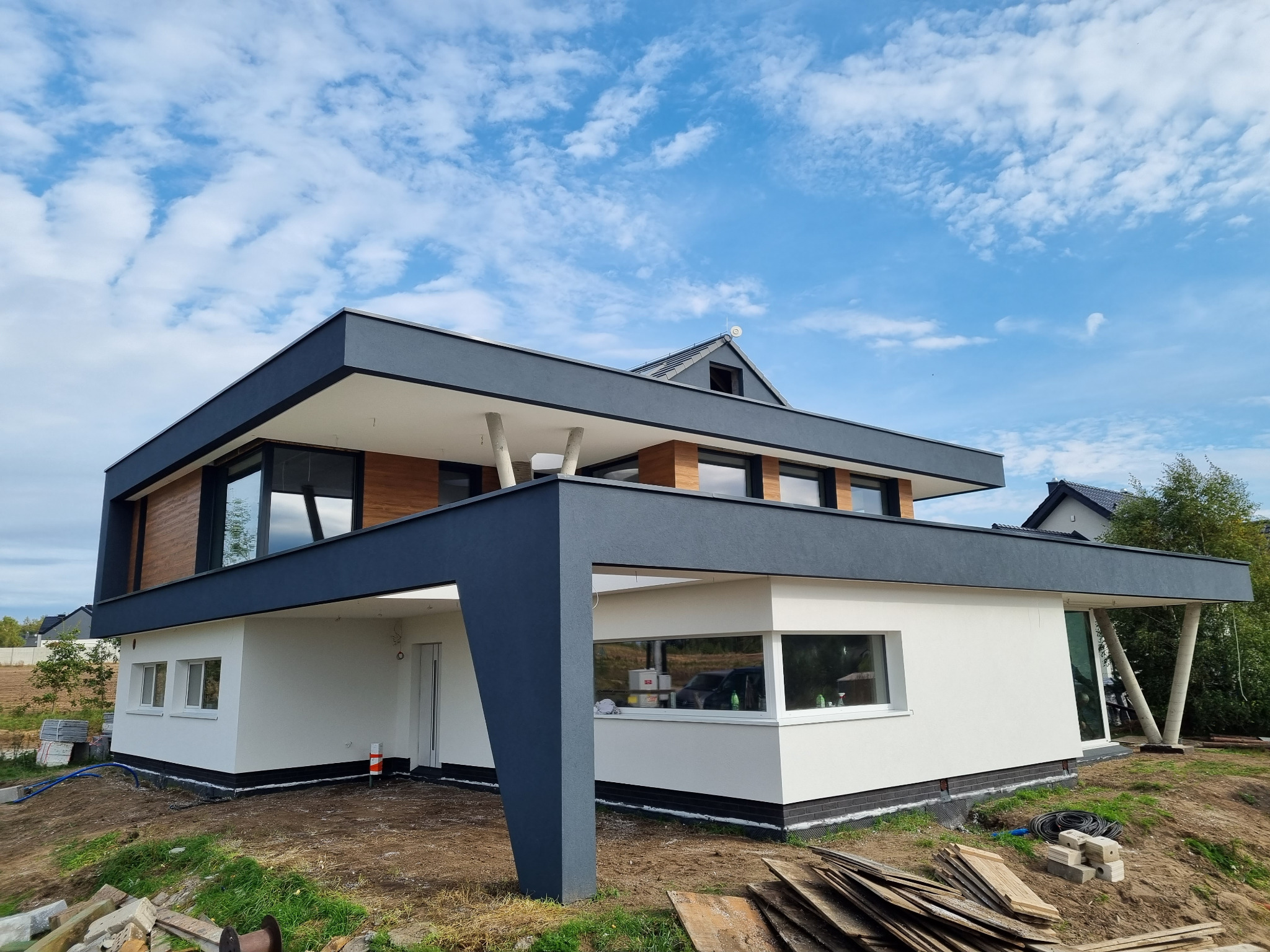 Nowoczesny dom dwukondygnacyjny z płaskim dachem, wykończony drewnem i szarą elewacją, widoczny z perspektywy gruntu, z elementami wykończeniowymi wokół fundamentów.