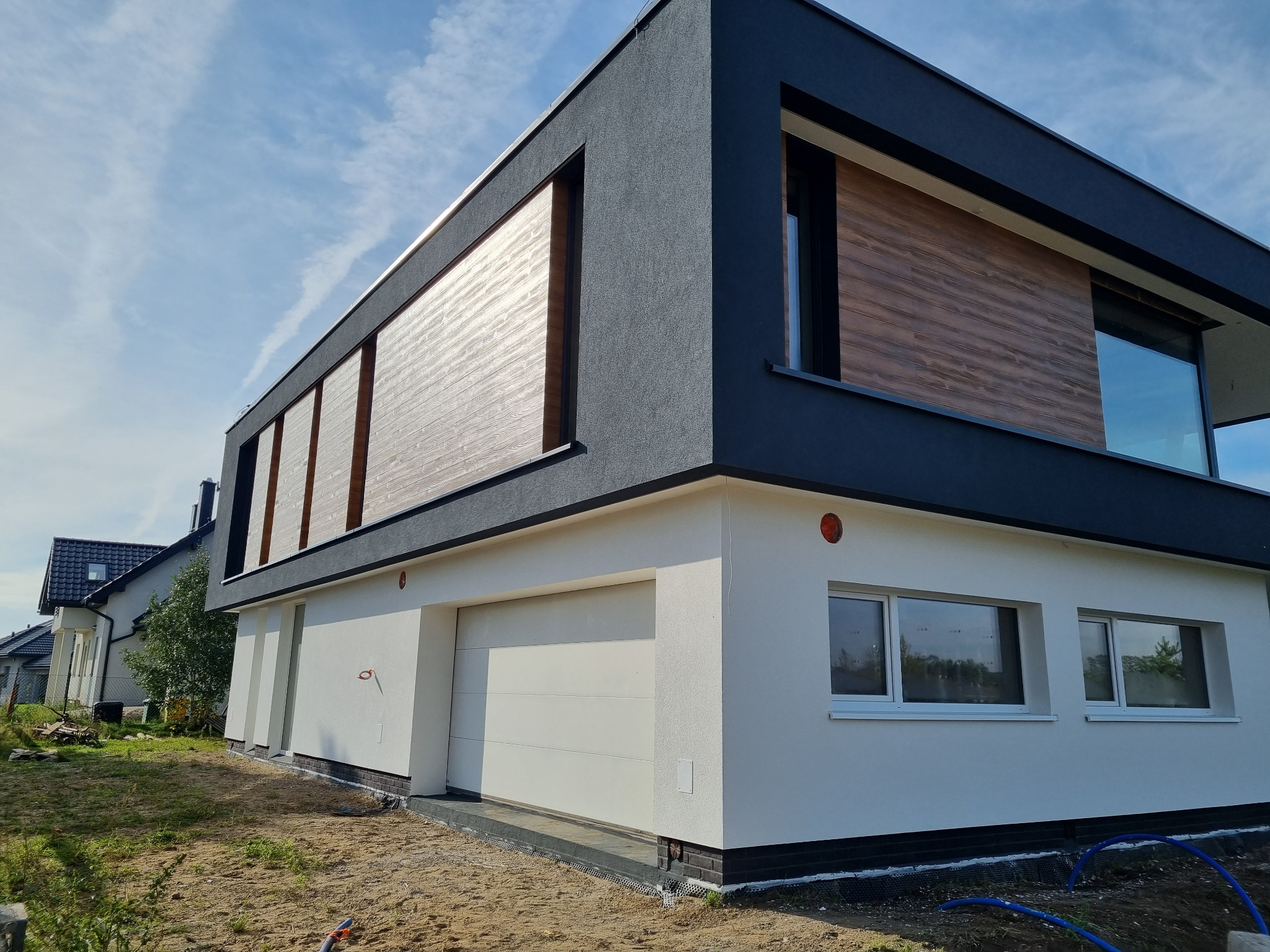 Nowoczesny dom jednorodzinny w Sierakowicach z elewacją w kolorze bieli i grafitu oraz drewnopodobnymi panelami, widok z zewnątrz na garaż i okna.