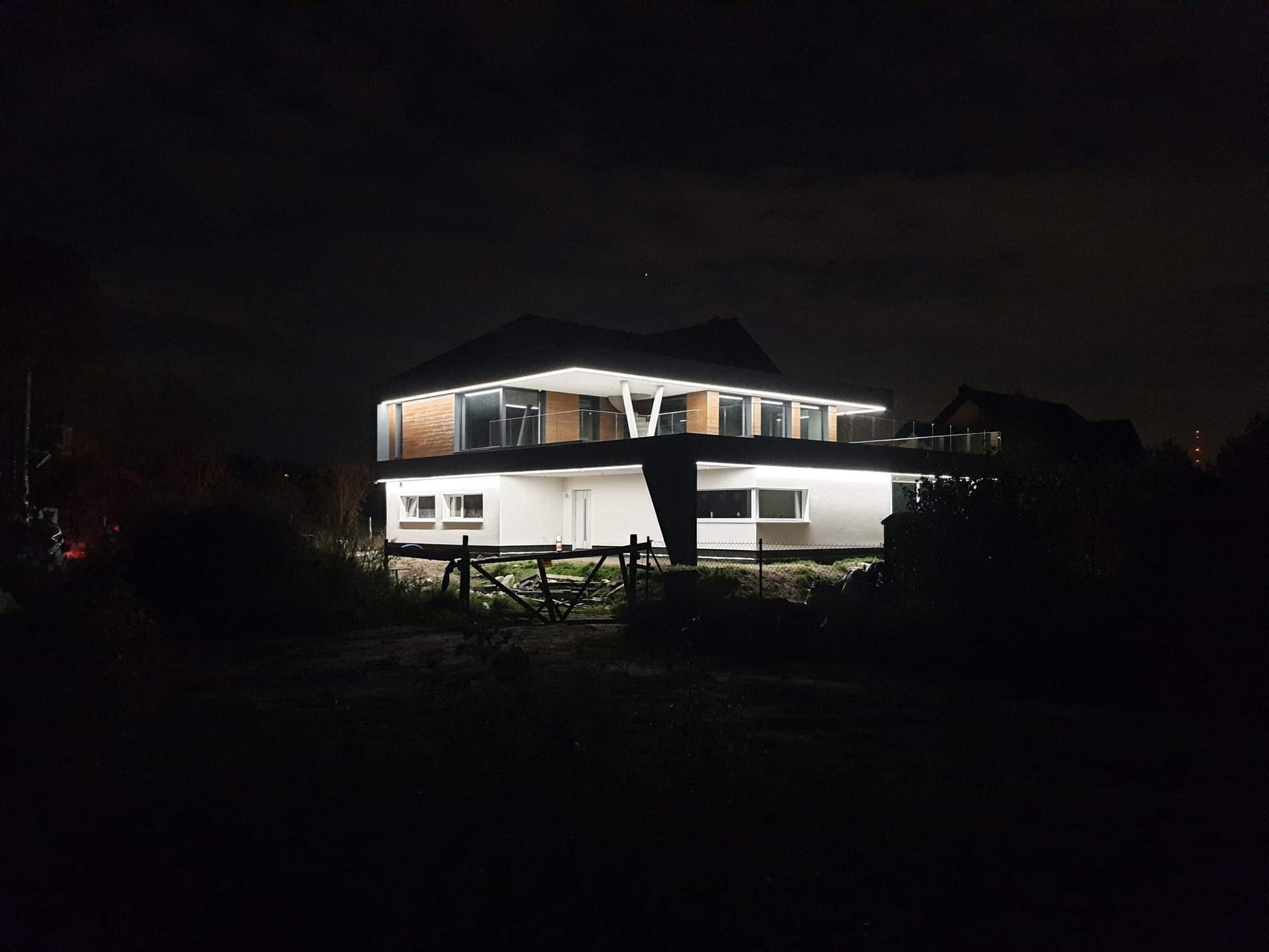 Dwupiętrowy dom z płaskim dachem, oświetlony nocą, z elementami drewnianej elewacji i białymi ścianami, widoczny z oddali przez niskie krzaki i prowizoryczne ogrodzenie.