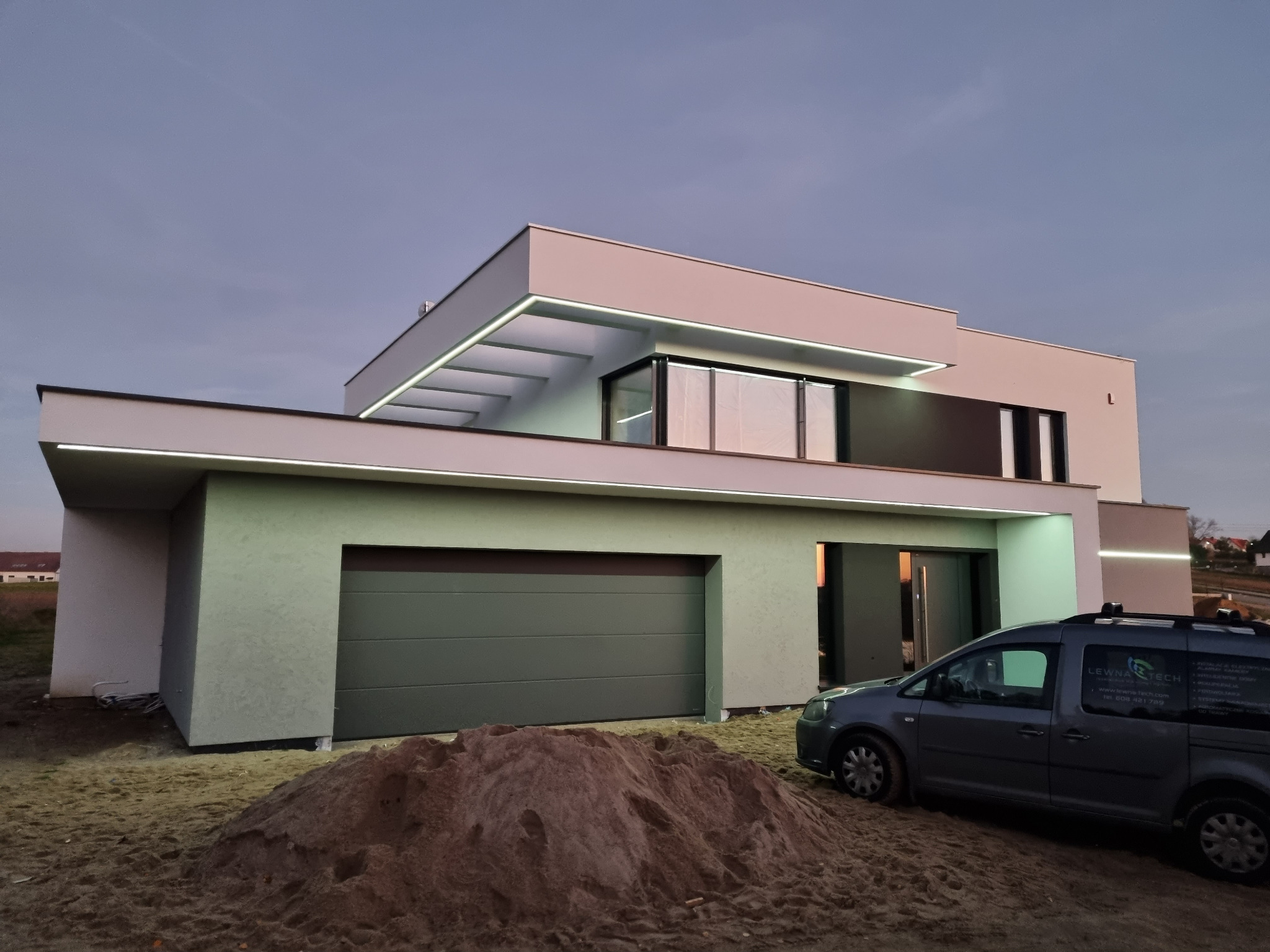 Nowoczesny dom jednorodzinny z płaskim dachem i oświetleniem LED elewacji, widoczny samochód dostawczy z logo firmy na tle sterty piasku.