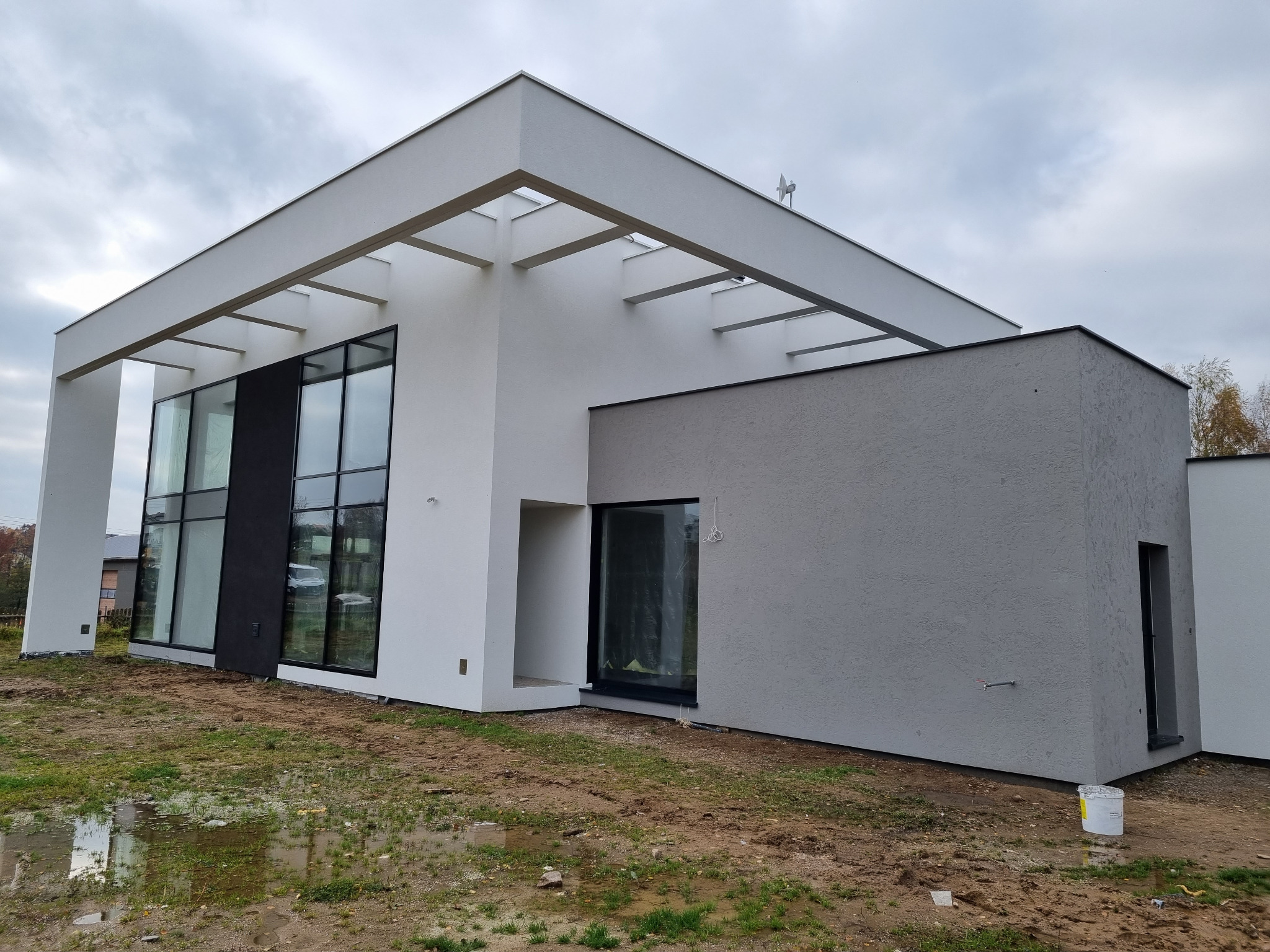 Nowoczesny dom z białą i szarą elewacją, dużymi oknami i charakterystyczną konstrukcją dachu, widoczny kubeł po farbie na trawniku.