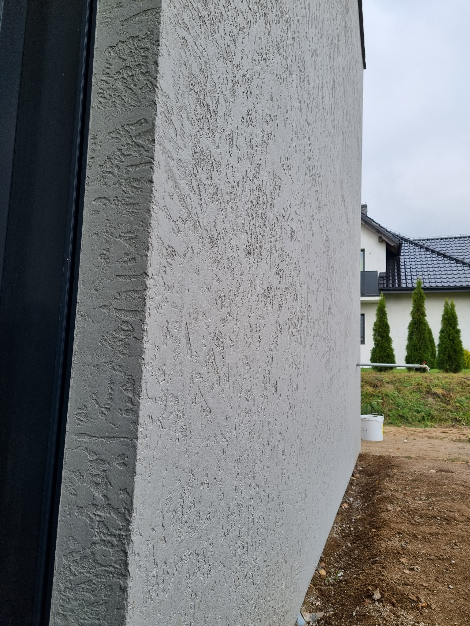 Fragment ściany budynku z szarą elewacją typu baranek, widok z boku, z elementami stolarki okiennej w tle i fragmentem ogrodu.