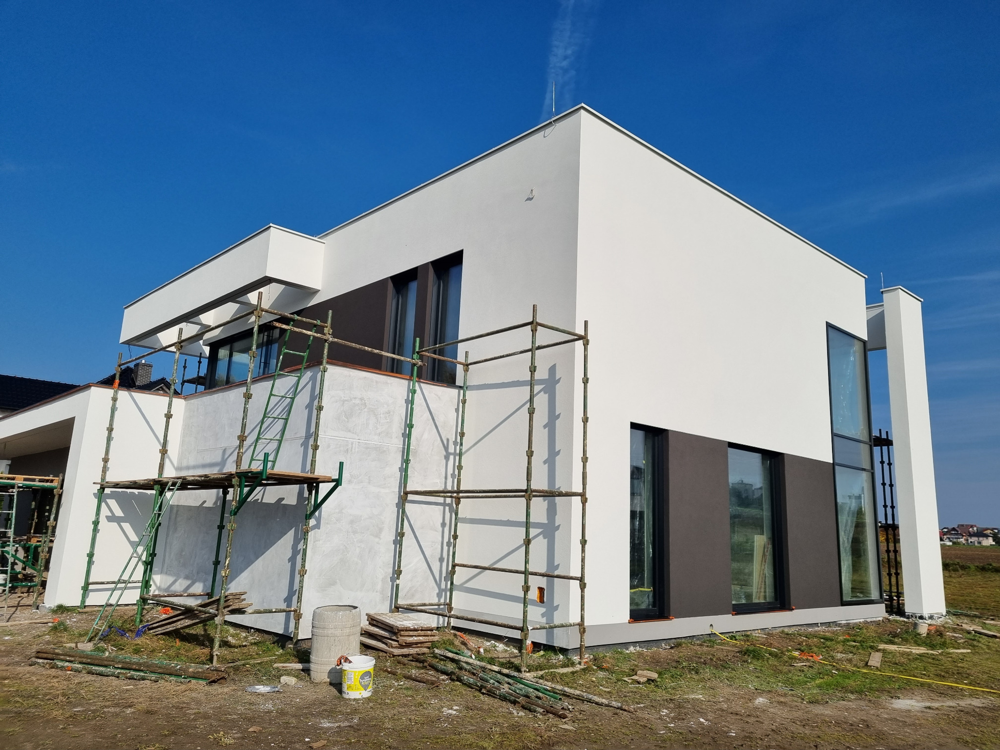 Nowoczesny dom jednorodzinny z białą i grafitową elewacją, w trakcie prac wykończeniowych, widoczne rusztowania i wiadro z farbą u podnóża ściany.