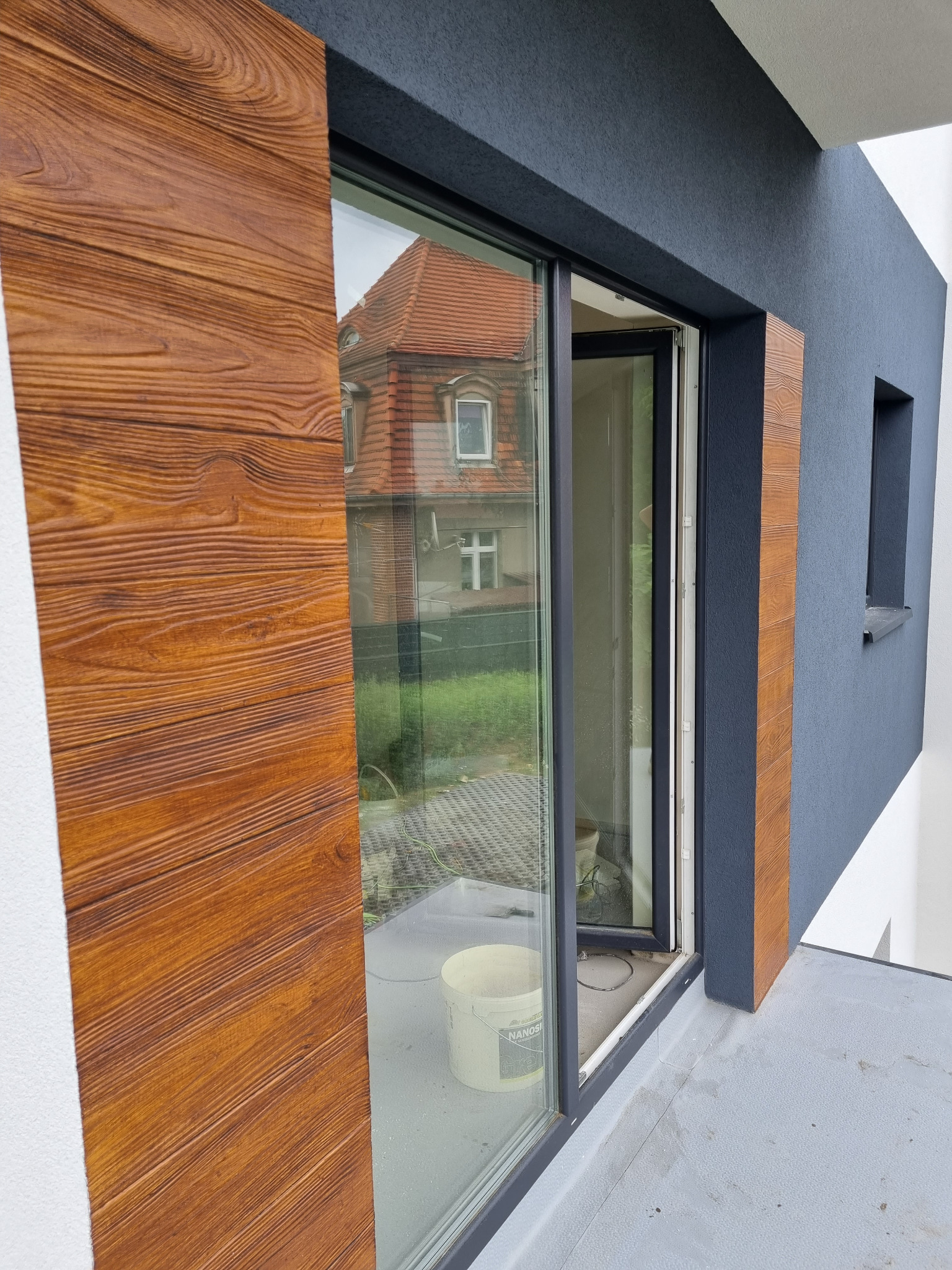 Nowoczesna elewacja domu z oknem, detale wykończenia w kolorze drewna, widoczna faktura tynku strukturalnego w odcieniach szarości i bieli, w odbiciu okna sąsiedni budynek z czerwonym dachem.