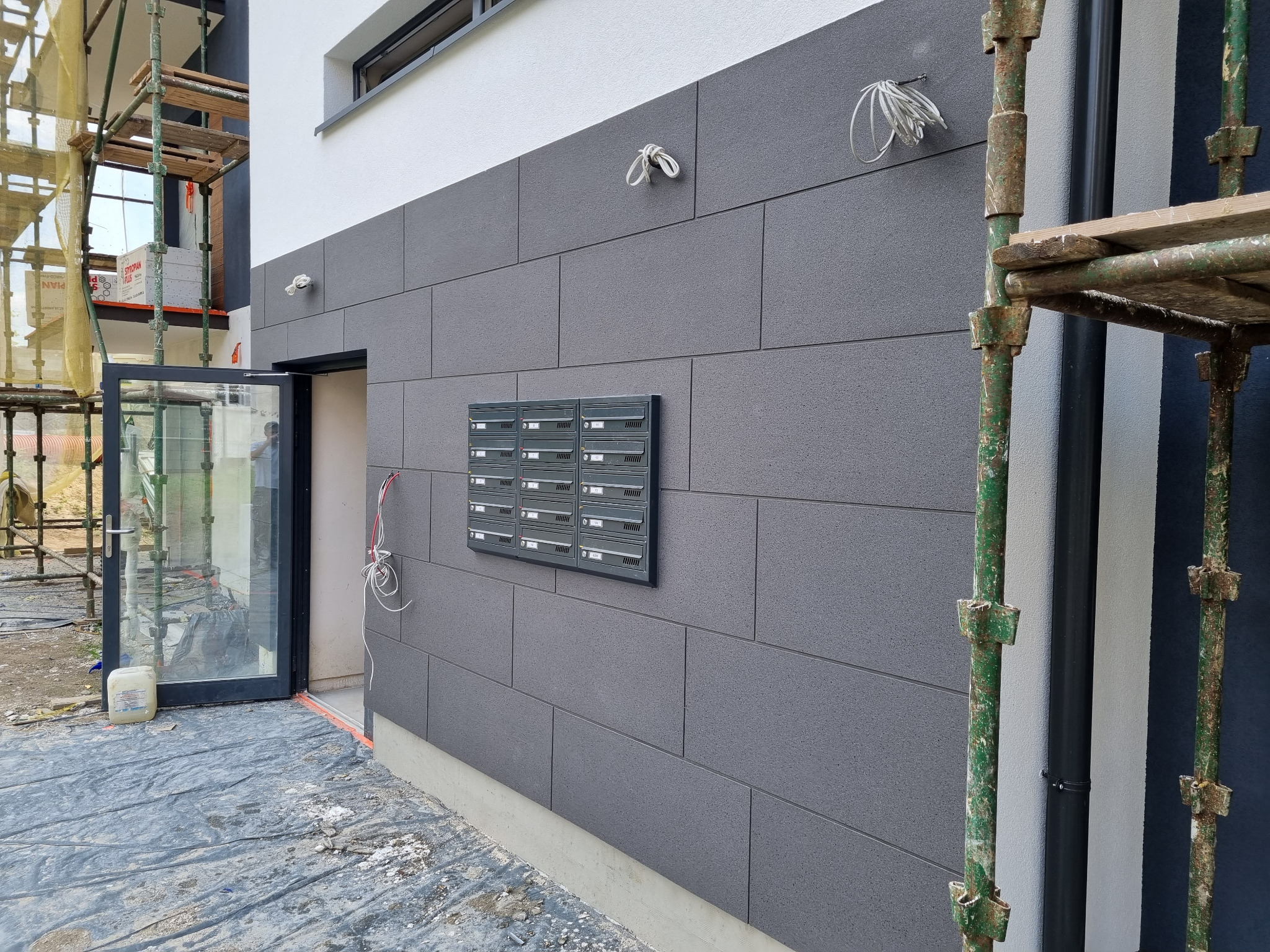 Szara elewacja budynku wielorodzinnego z zamontowanymi skrzynkami pocztowymi, widoczne rusztowania i przewody elektryczne wystające ze ściany.