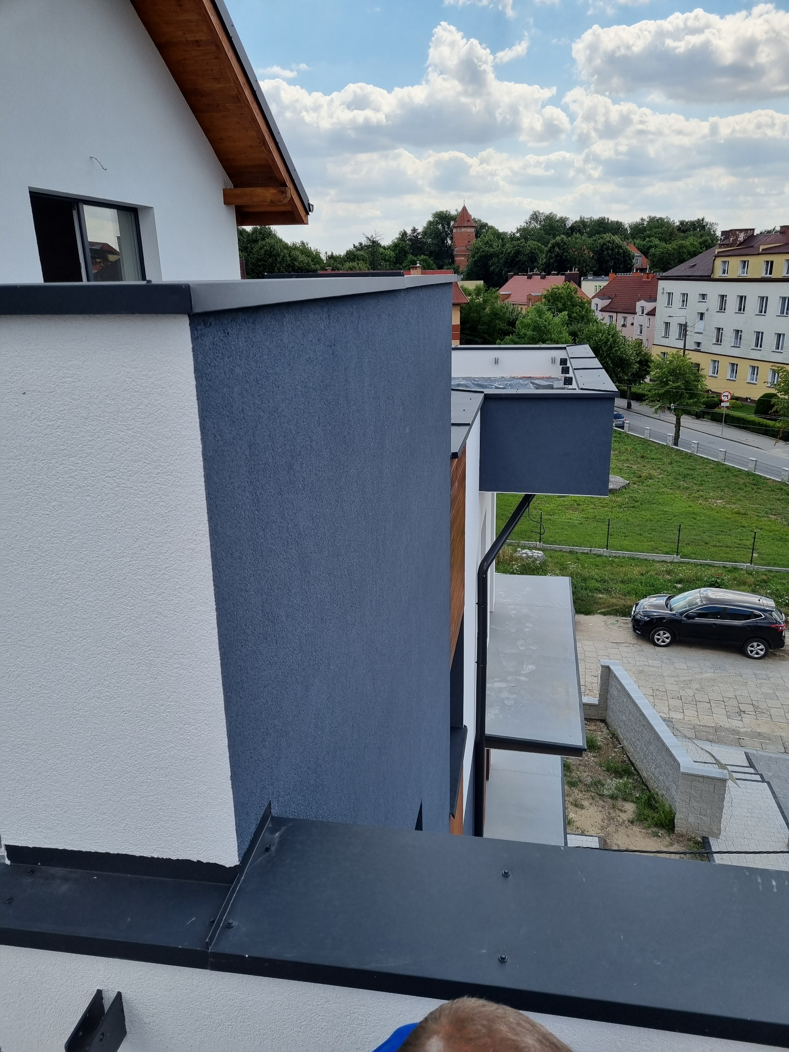 Nowoczesna elewacja budynku z wyraźnym podziałem na białą i grafitową część, widok z góry, w tle zabudowa miejska i błękitne niebo z chmurami.