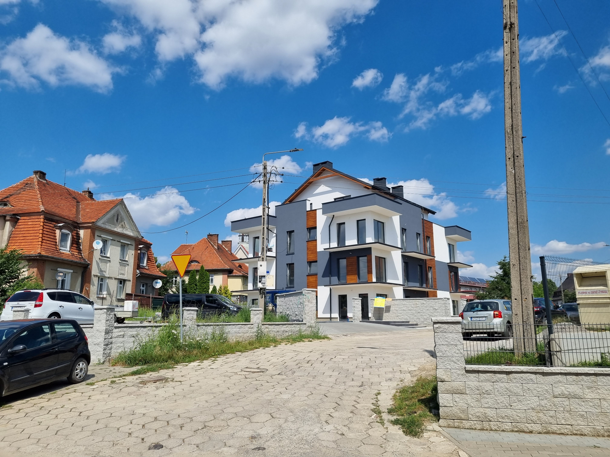 Nowoczesny budynek wielorodzinny z elewacją w kolorach bieli i szarości, z elementami drewnopodobnymi, widoczny z perspektywy ulicy, z brukowaną drogą i słupem energetycznym po prawej stronie.