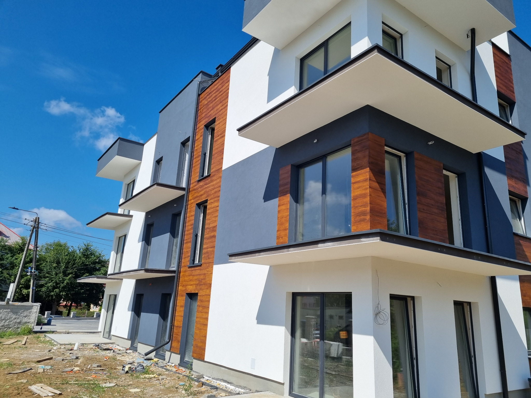 Nowoczesny budynek wielorodzinny z elewacją w kolorach białym, szarym i drewnopodobnym, z widocznymi balkonami i dużymi oknami, sfotografowany w słoneczny dzień na tle błękitnego nieba,...