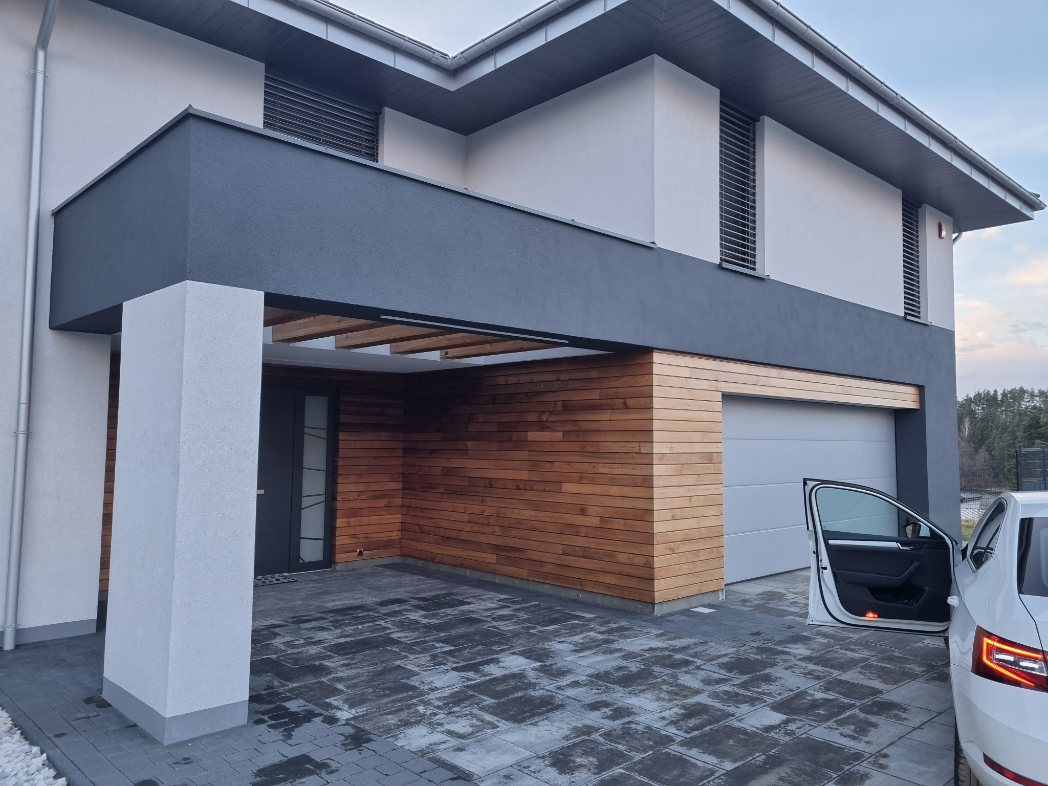 Nowoczesny dom jednorodzinny z elementami drewnianej elewacji i szarą bryłą, widok z podjazdu wyłożonego kostką, z otwartymi drzwiami samochodu osobowego.