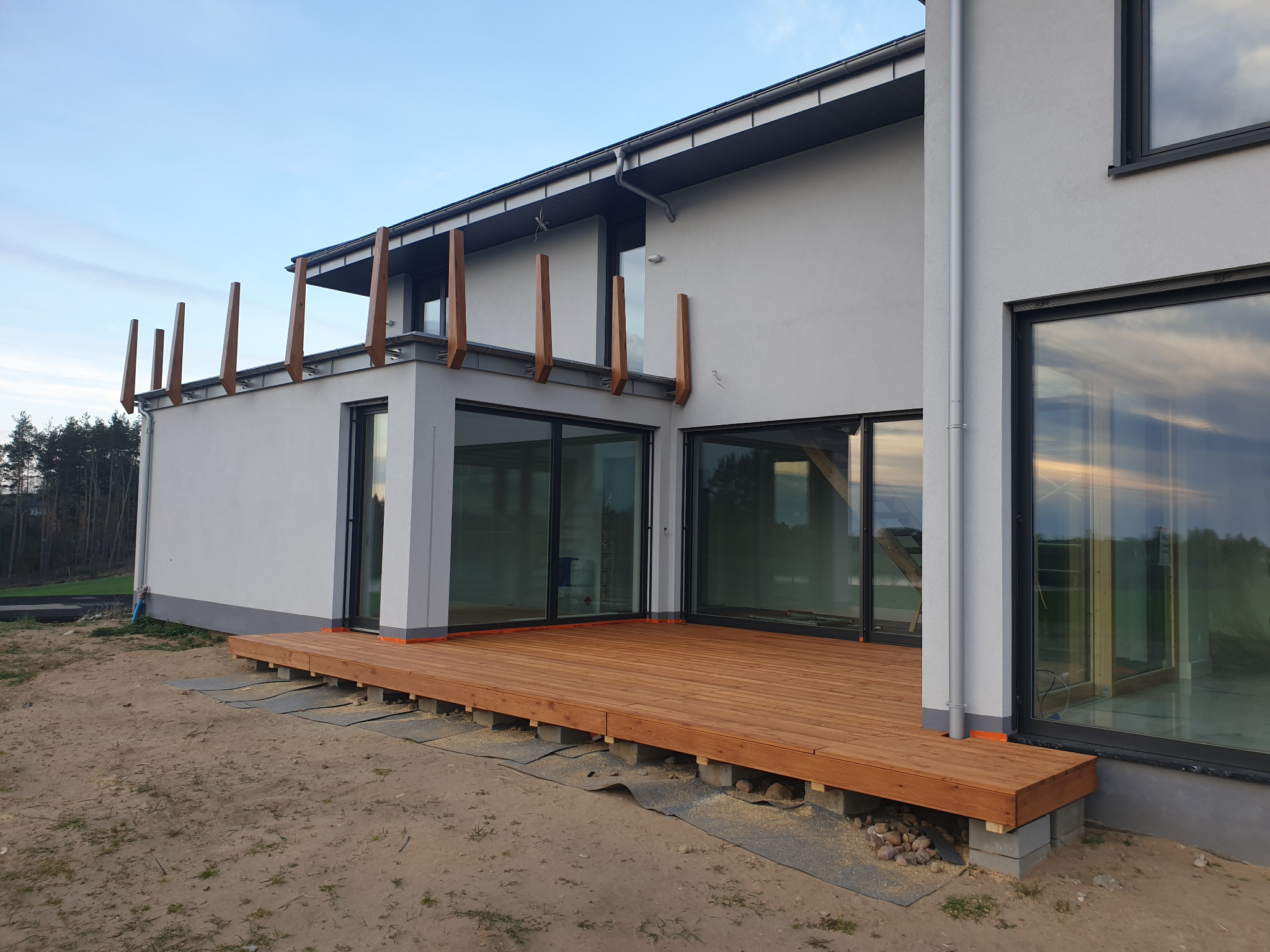 Nowoczesny dom z szarą elewacją i drewnianym tarasem na betonowych bloczkach, widoczne duże okna z odbiciem nieba, elementy drewniane na balkonie.