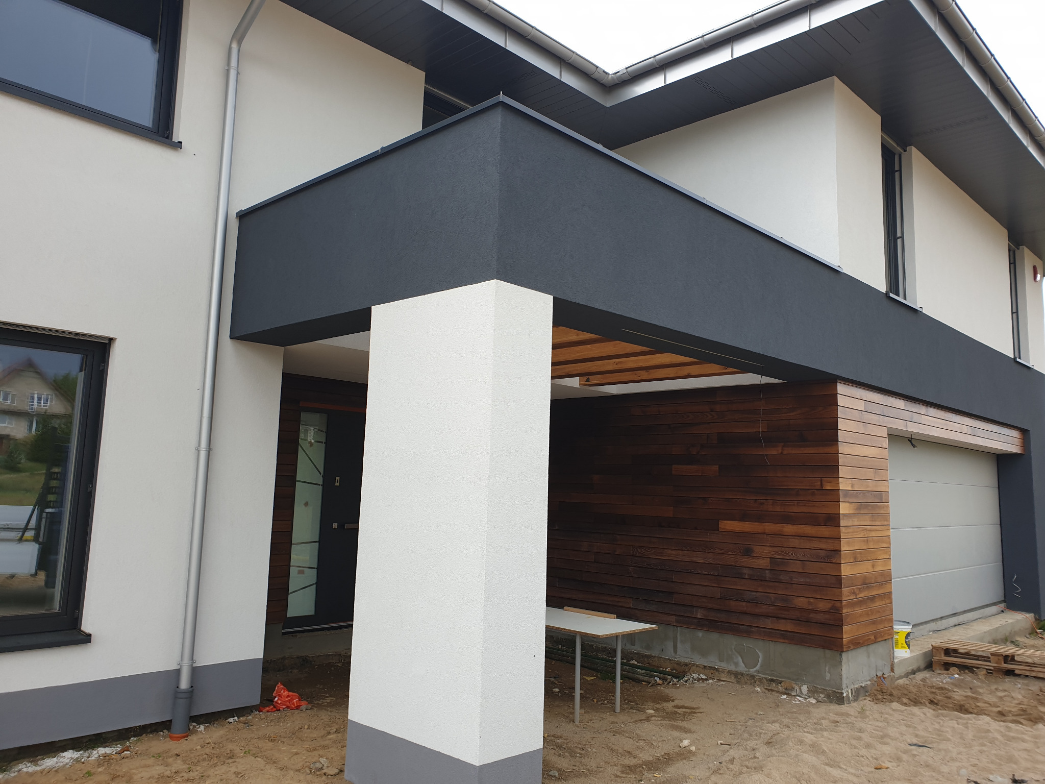 Nowoczesny dom jednorodzinny z elewacją w kolorach bieli i grafitu, z drewnianą okładziną garażu, widoczna kolumna wejściowa i fragment terenu budowy.