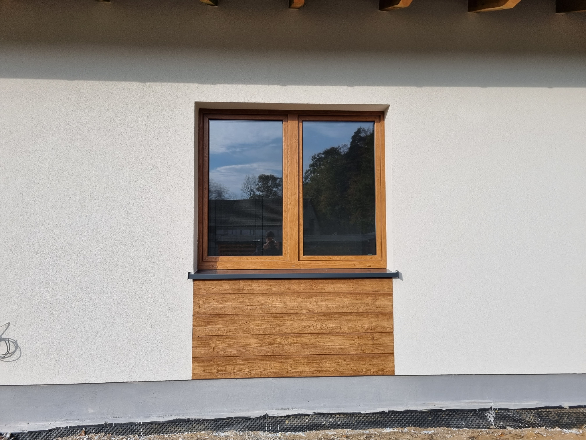Fragment elewacji budynku z oknem w drewnianej ramie i dekoracyjnym panelem imitującym drewno pod oknem, widoczna faktura tynku.