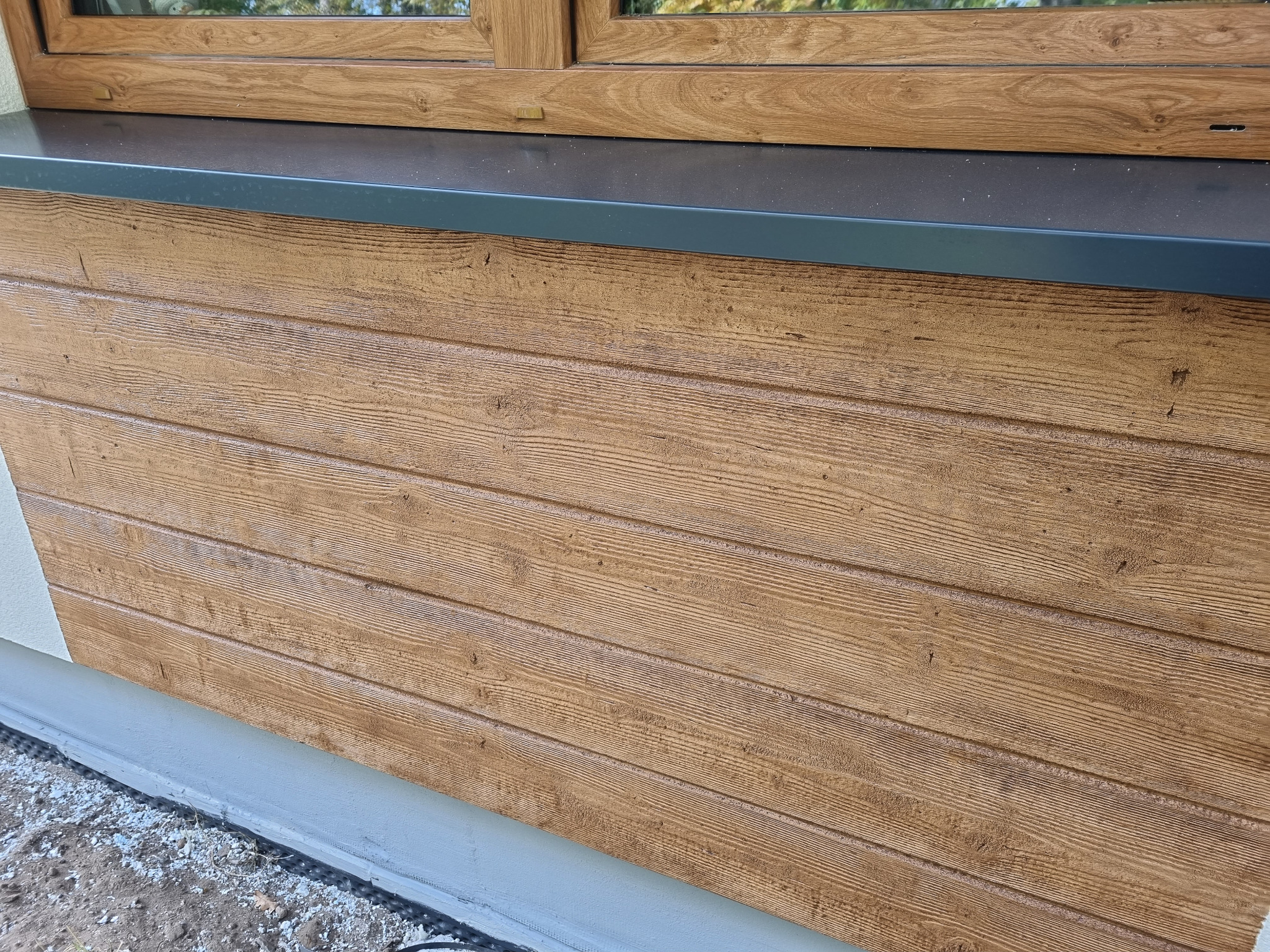 Fragment elewacji budynku z drewnopodobną okładziną poziomą, widoczne okno z brązową ramą i szary parapet oraz fragment izolacji przy fundamencie.