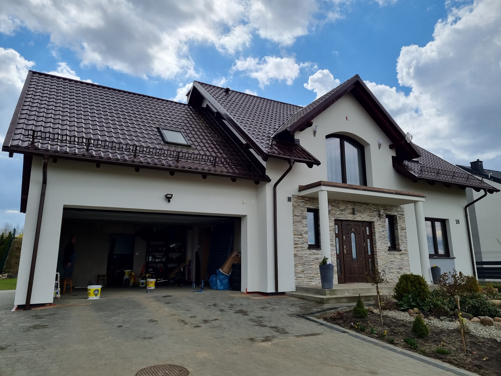 Nowoczesny dom jednorodzinny z kremową elewacją, brązowym dachem z dachówki, kamiennym wykończeniem wejścia i otwartym garażem, w którym widać puszki z farbą.