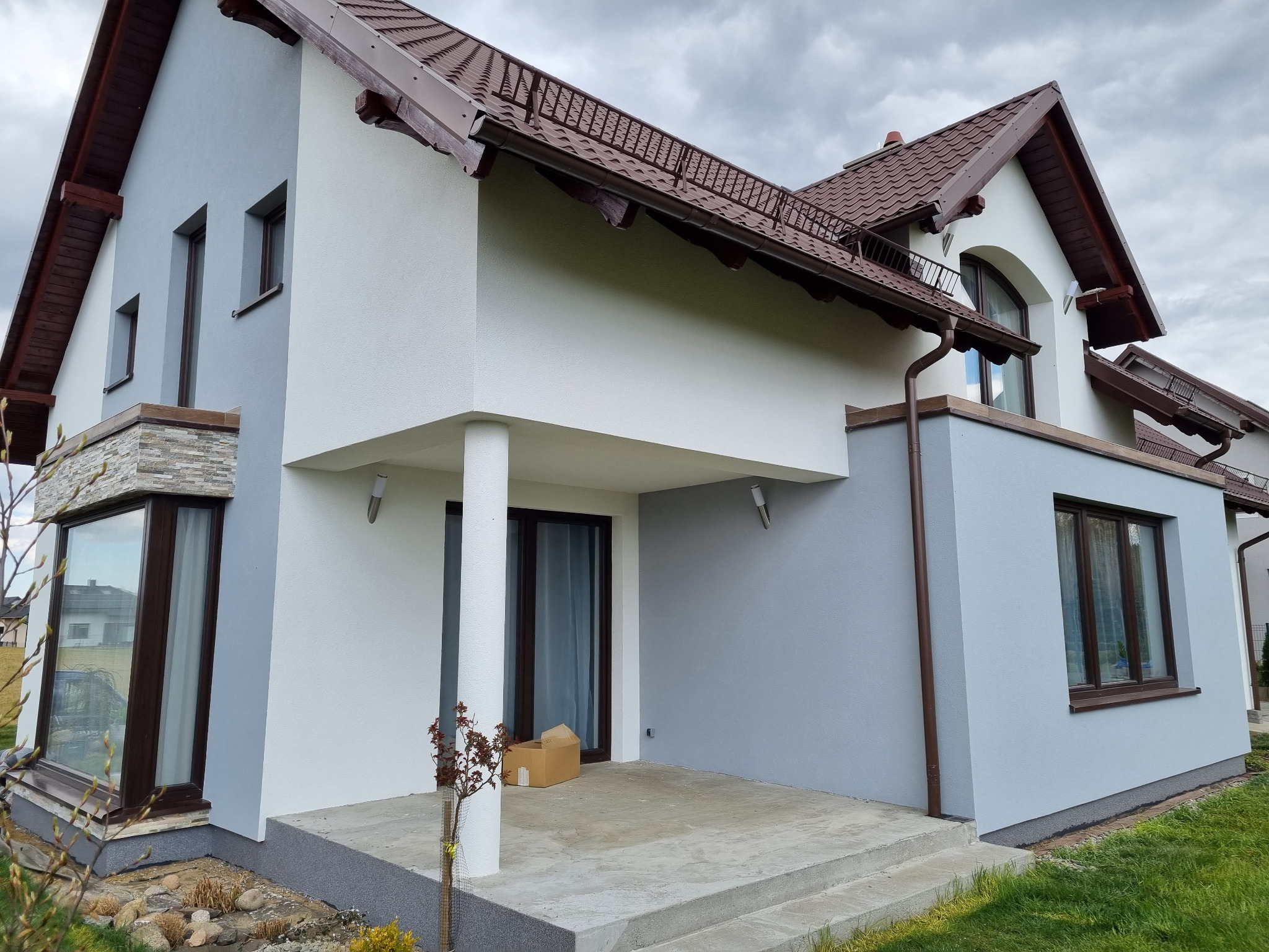Nowoczesny dom jednorodzinny z elewacją w odcieniach szarości i bieli, brązowy dach z rynną, widoczny fragment tarasu z kartonami, w tle pochmurne niebo.