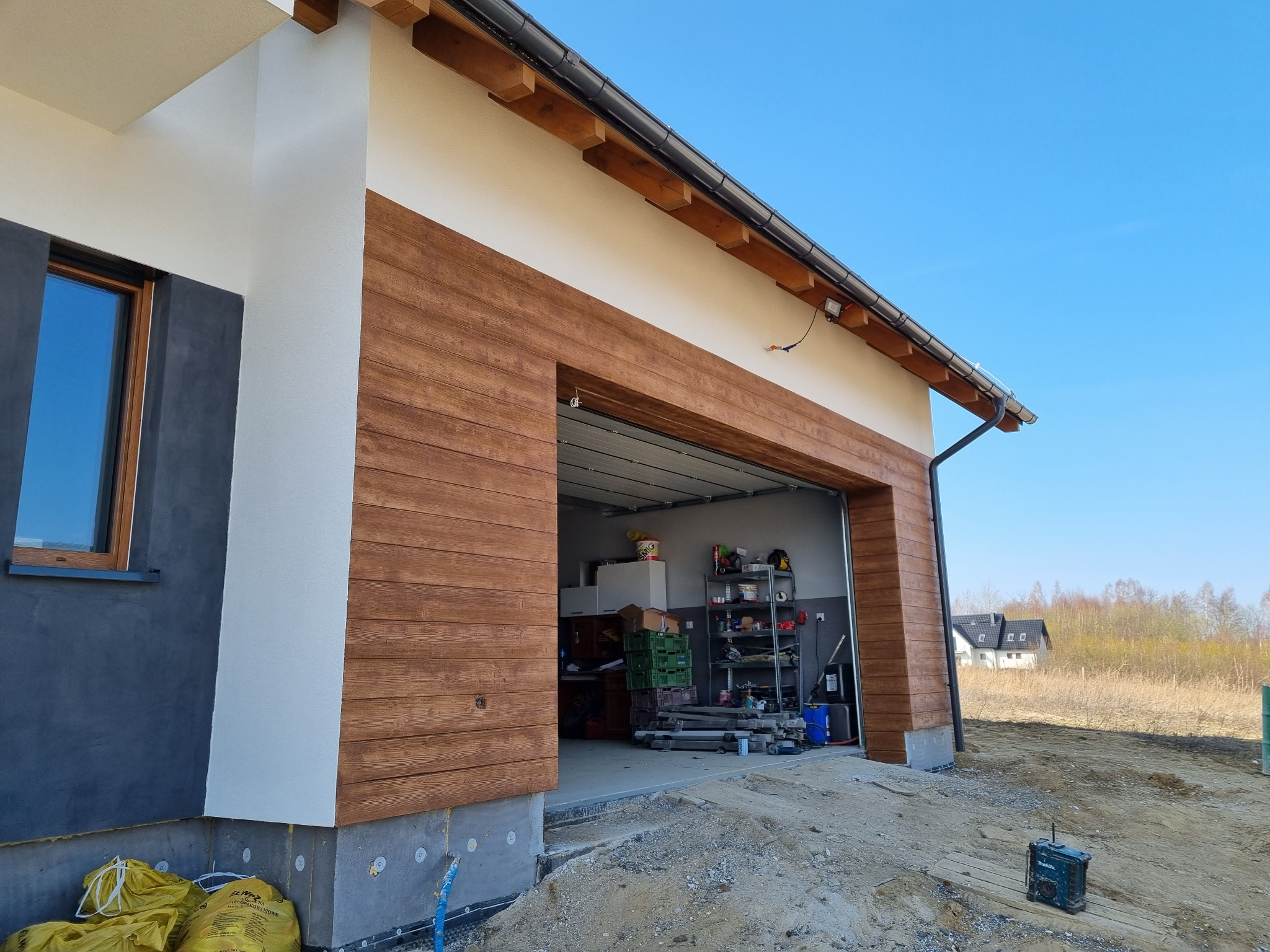 Nowoczesny garaż z elewacją imitującą drewno, widoczne worki z materiałem izolacyjnym i narzędzia przed budynkiem w trakcie prac wykończeniowych.
