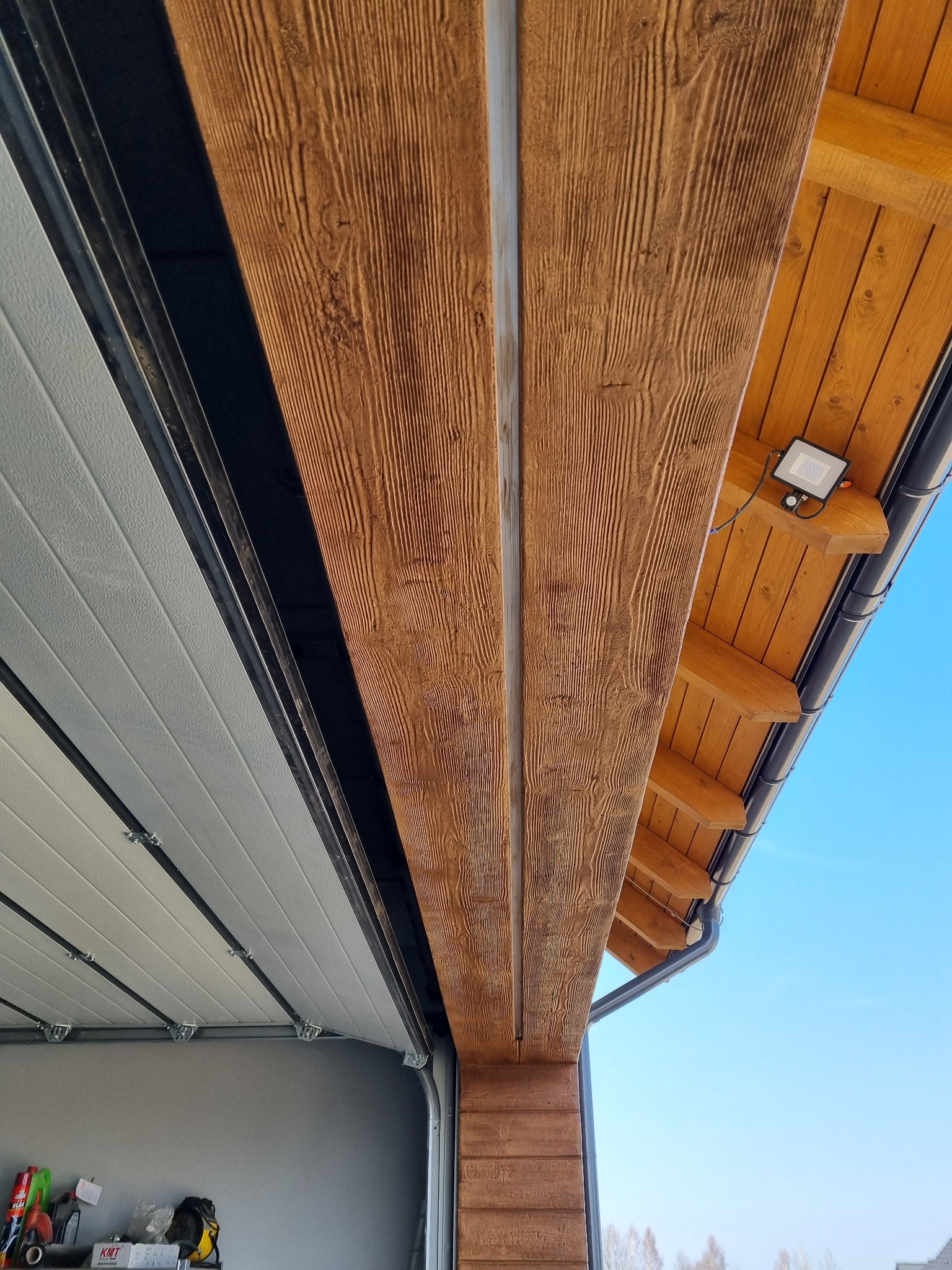 Fragment elewacji budynku z widocznym oświetleniem zewnętrznym, rynnami i otwartą bramą garażową. Elewacja imituje drewno.