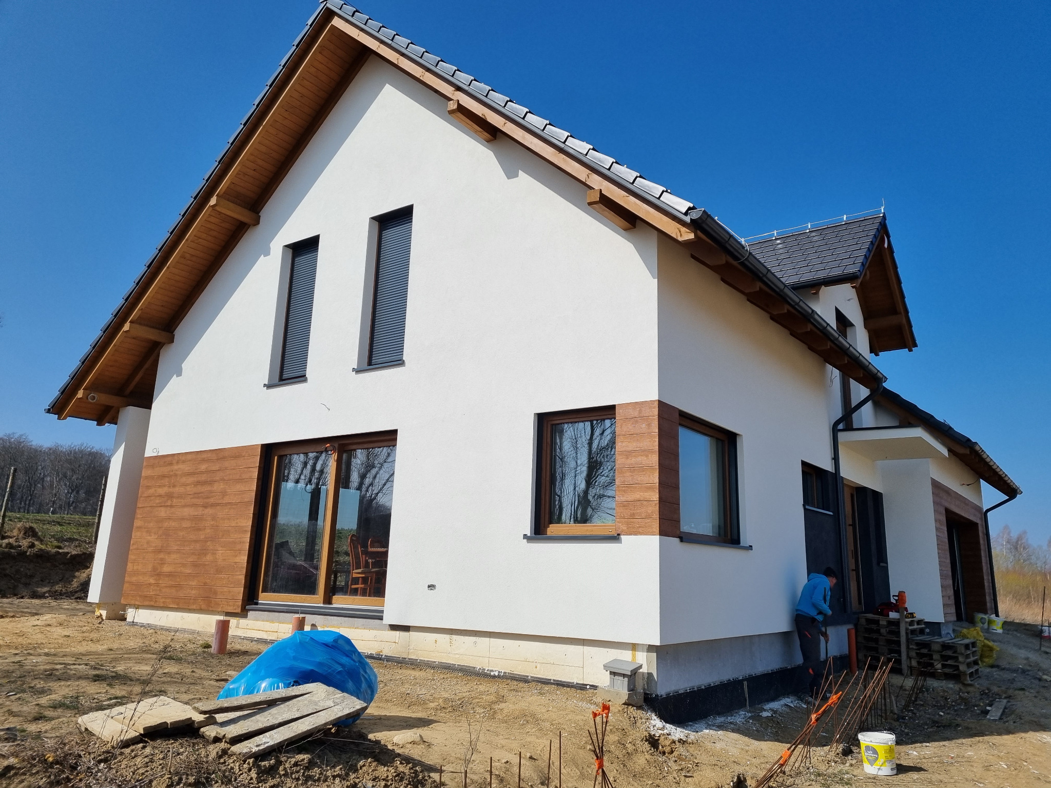 Nowoczesny dom z białą elewacją i drewnianymi akcentami, widoczny fragment izolacji fundamentów oraz pracownik w trakcie wykonywania prac wykończeniowych przy wejściu.