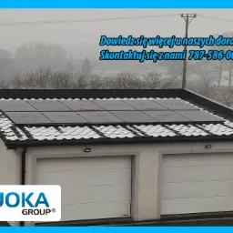 Panele fotowoltaiczne na dachu garażu dwustanowiskowego pokrytego śniegiem, logo firmy JOKA GROUP w lewym dolnym rogu, zima w tle.