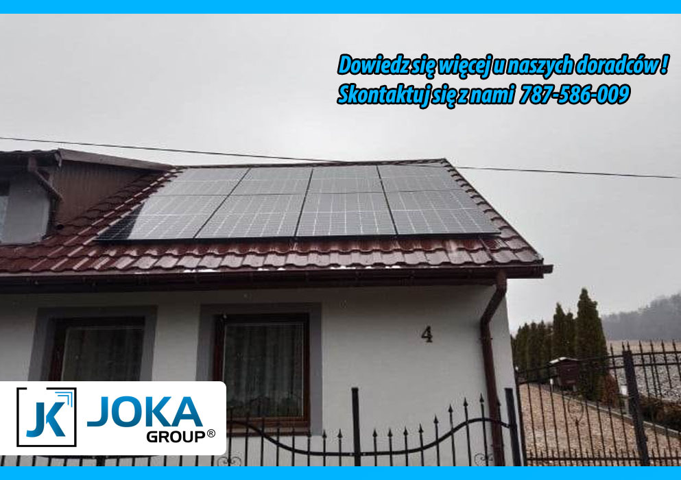 Panele słoneczne na dachu domu z brązową dachówką, numer domu '4' widoczny na elewacji, logo firmy JOKA GROUP w lewym dolnym rogu, reklama firmy z numerem telefonu w górnej części zdjęcia.
