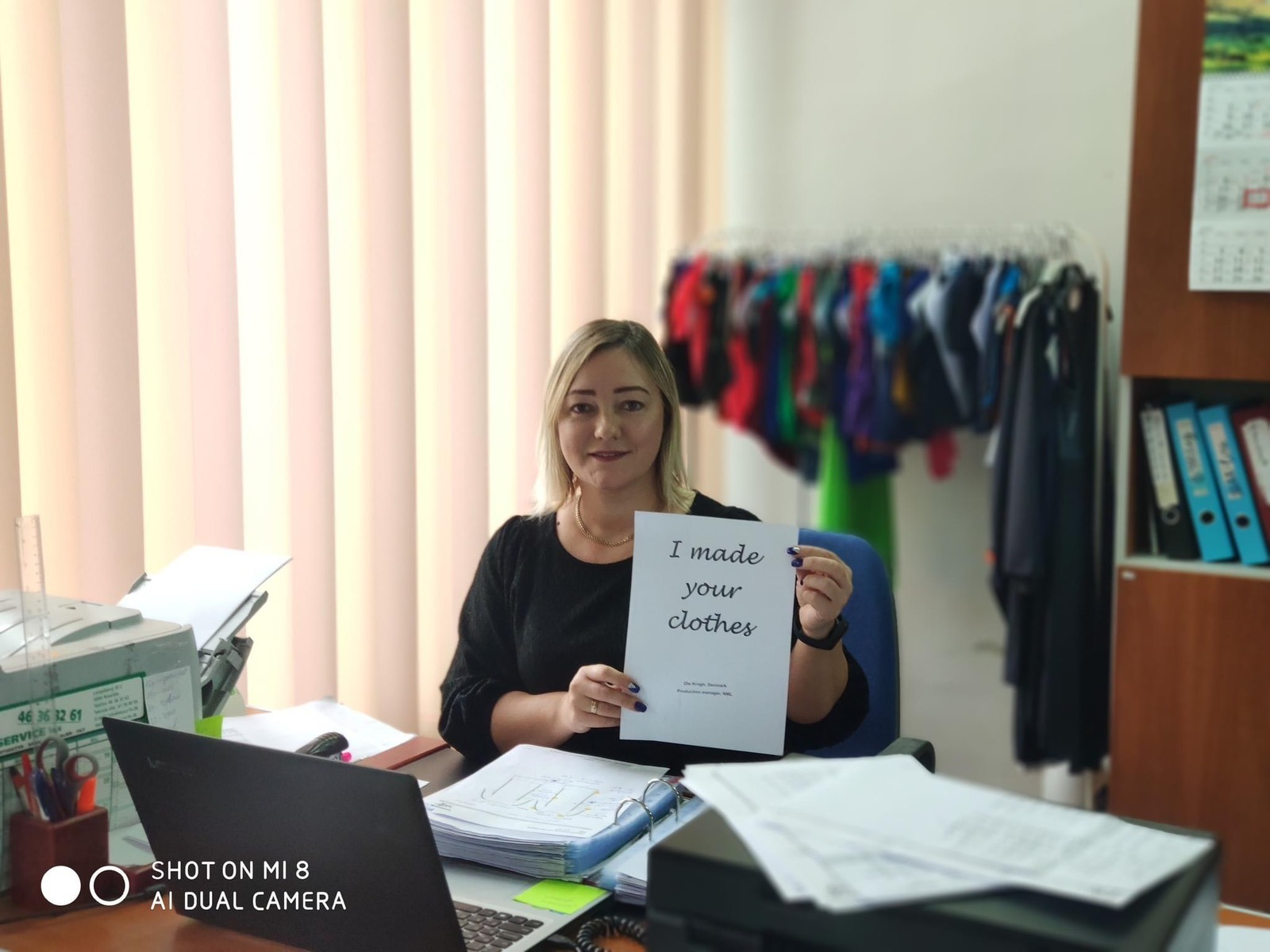 Kobieta w biurze trzyma kartkę z napisem 'I made your clothes', w tle wieszak z ubraniami i biurko z dokumentami.