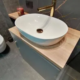 Nowoczesna łazienka z owalną białą umywalką nablatową, złotą baterią, drewnianym blatem i szarą szafką pod umywalką na tle ściany z ciemnego marmuru.