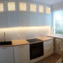 Jasna kuchnia z białymi szafkami, drewnianym blatem i złotą armaturą, oświetlona listwą LED pod szafkami wiszącymi, z widocznym oknem z żaluzjami i fragmentem grzejnika.