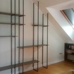 Regał z drewnianymi p&oacute;łkami i czarnymi metalowymi wspornikami, zamontowany na białej ścianie w pokoju na poddaszu z drewnianą podłogą i oknami dachowymi.