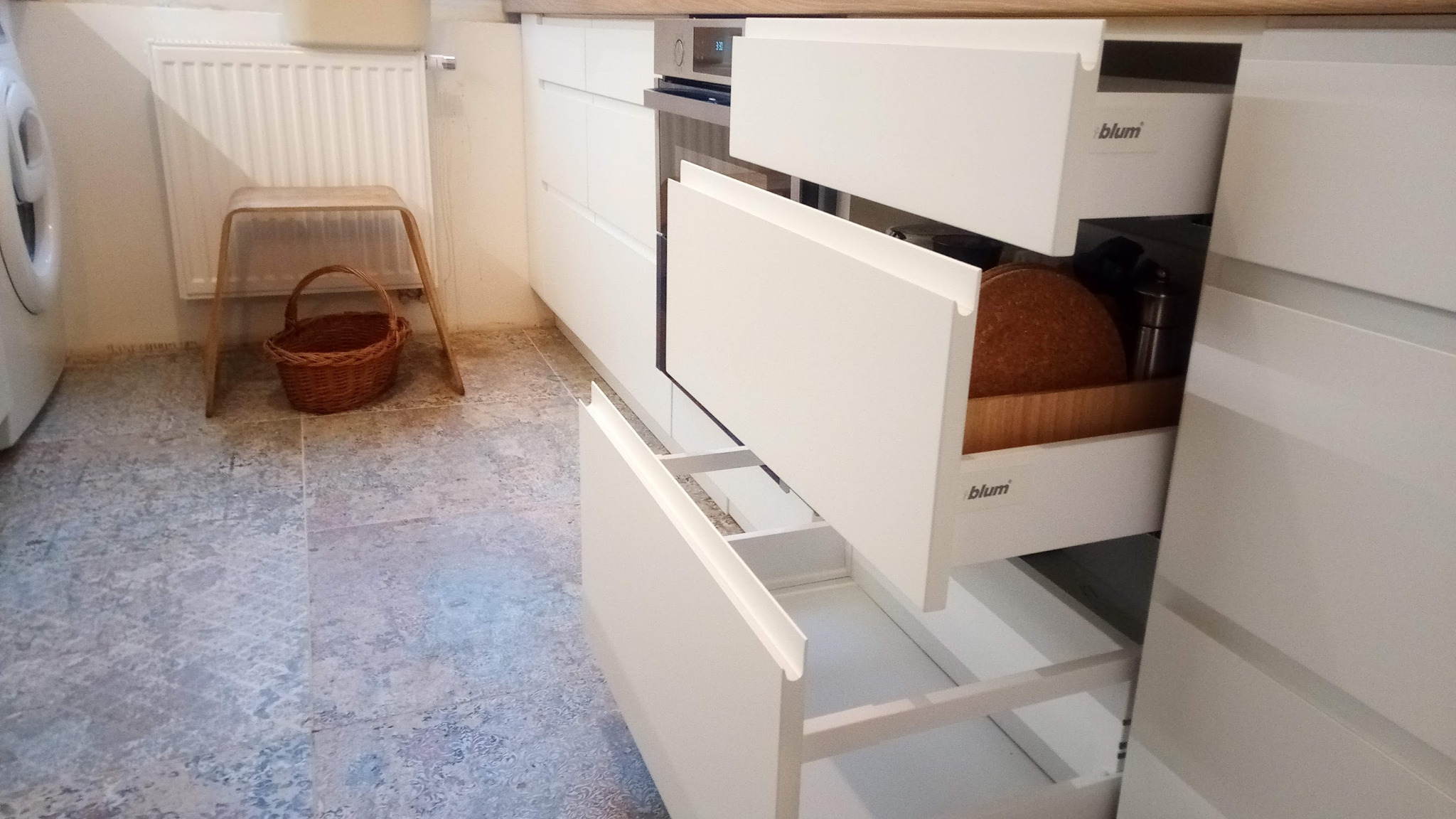 Biała zabudowa kuchenna z otwartymi szufladami, widoczne akcesoria kuchenne wewnątrz, na podłodze wzorzyste płytki, w tle pralka i drewniany stołek z koszykiem.