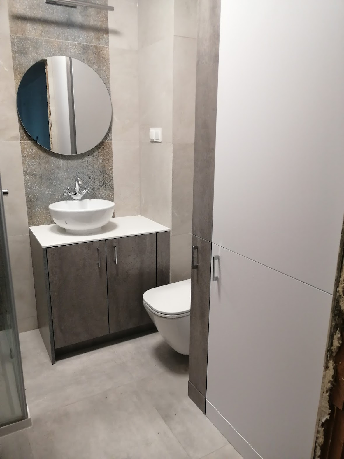 Nowoczesna łazienka z okrągłym lustrem, umywalką nablatową na szafce w kolorze szarości i białą szafą w zabudowie, uzupełniona wiszącą toaletą.