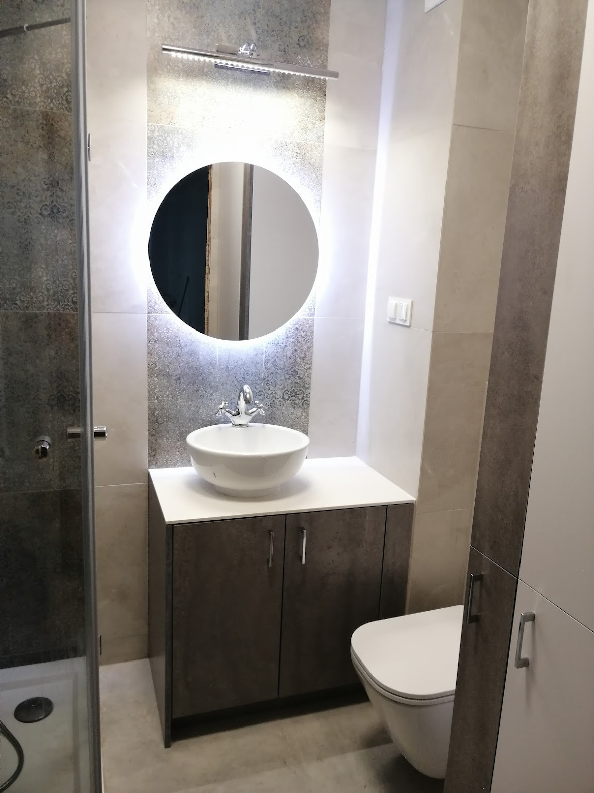 Nowoczesna łazienka z okrągłym lustrem podświetlanym LED, umywalką nablatową na szafce z ciemnymi frontami i kabiną prysznicową z szarymi płytkami.