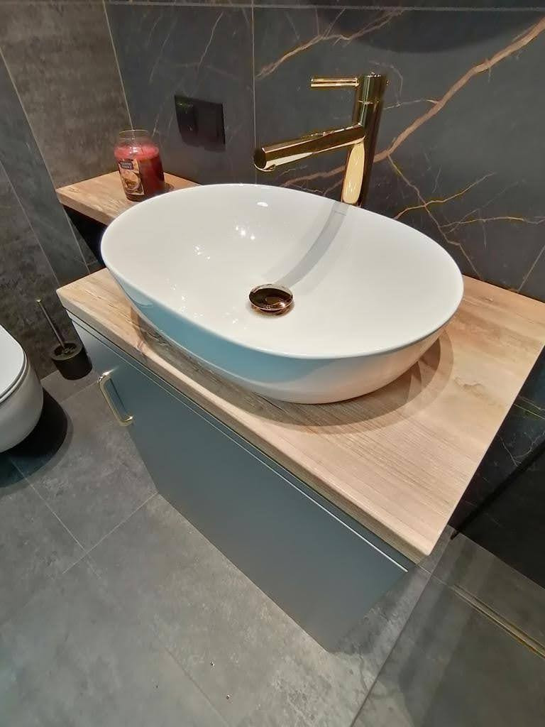 Nowoczesna łazienka z owalną białą umywalką nablatową, złotą baterią, drewnianym blatem i szarą szafką pod umywalką na tle ściany z ciemnego marmuru.