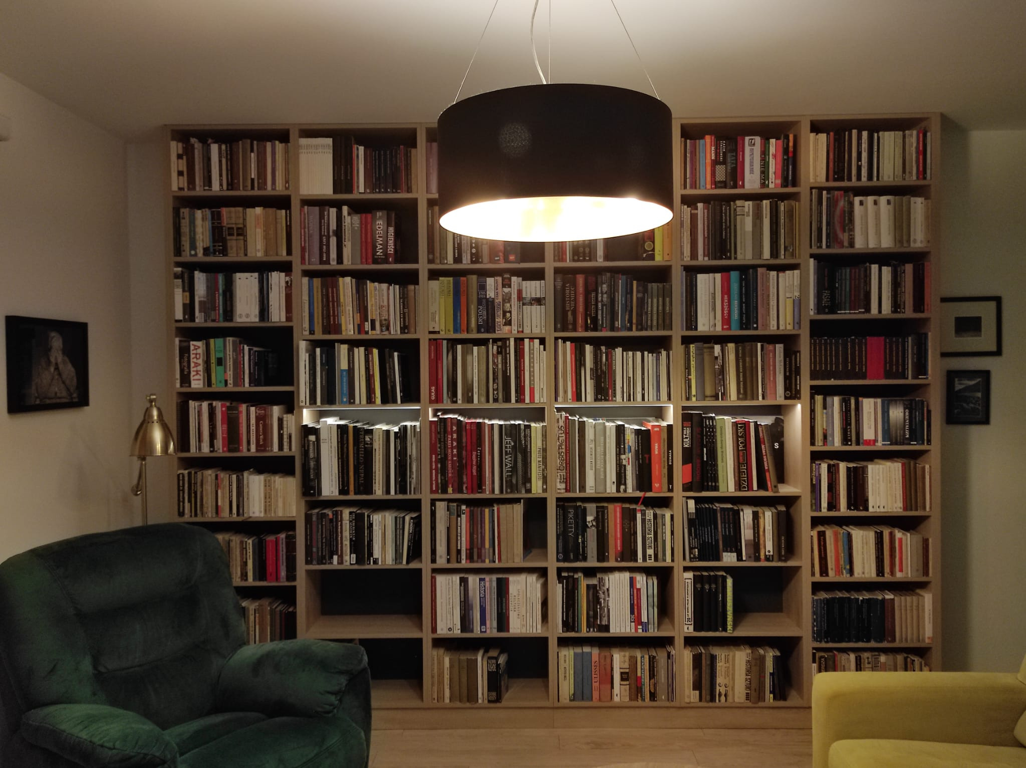 Duża, drewniana biblioteczka na wymiar wypełniająca całą ścianę we Wrocławiu, oświetlona od góry wiszącą lampą i listwami LED, z zielonym fotelem i żółtą sofą w tle.
