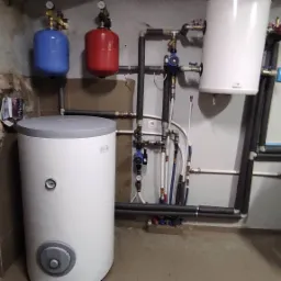 Bojler z szarym wykończeniem, podłączony do instalacji grzewczej z czerwonym i niebieskim zbiornikiem wyrównawczym, widoczne rury i zawory w pomieszczeniu technicznym.