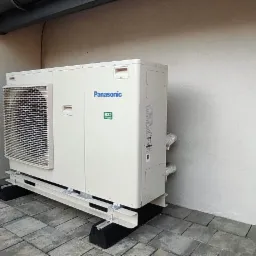 Zewnętrzna jednostka pompy ciepła Panasonic zamontowana na podwyższeniu z metalową ramą i gumowymi podkładkami, ustawiona na kostce brukowej przy jasnej ścianie budynku.