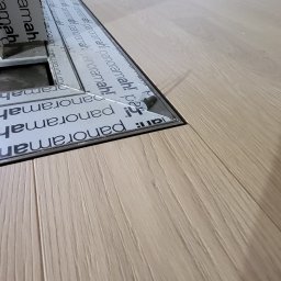 Ecobat Sarl - Fragment podłogi z jasnych paneli z widocznym metalowym profilem podłogowym z nadrukiem 'panoramah!', otaczającym otwór w podłodze.