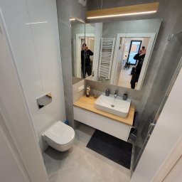 Ecobat Sarl - Nowoczesna łazienka z szarą płytką imitującą marmur, podwieszana toaleta z chromowanym przyciskiem spłukiwania, umywalka nablatowa na drewnianym blacie i lustro z oświetleniem LED, widoczny...