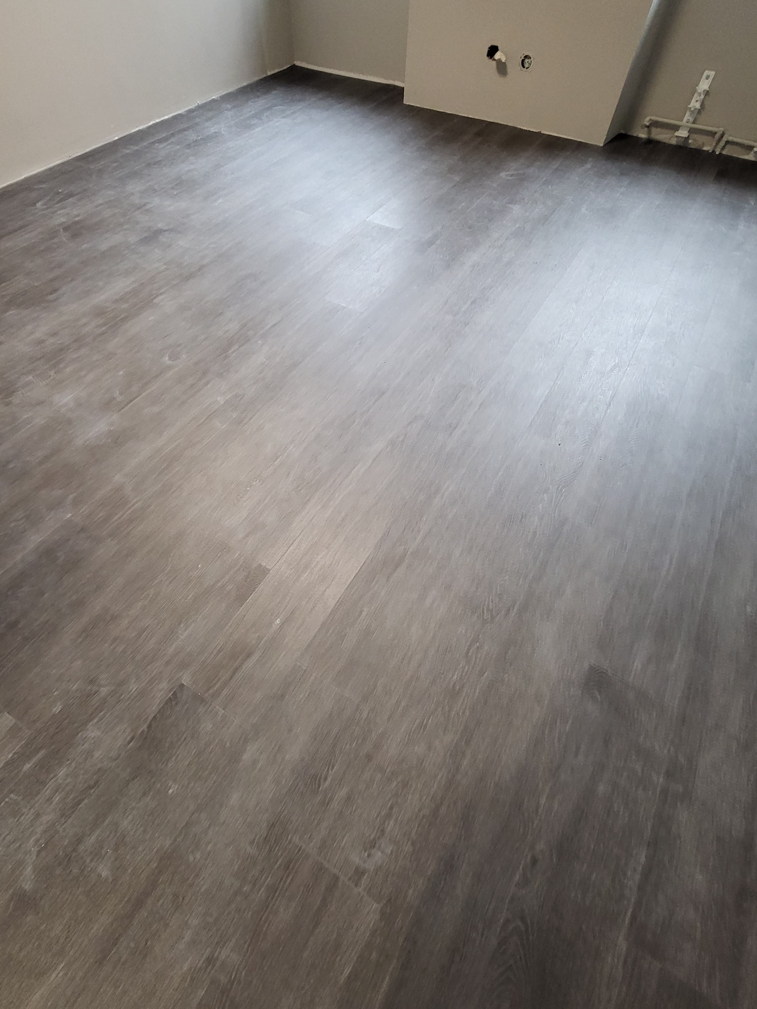 Nowo położona, szara podłoga panelowa w pustym pomieszczeniu z widocznymi rurami instalacji grzewczej przy ścianie.