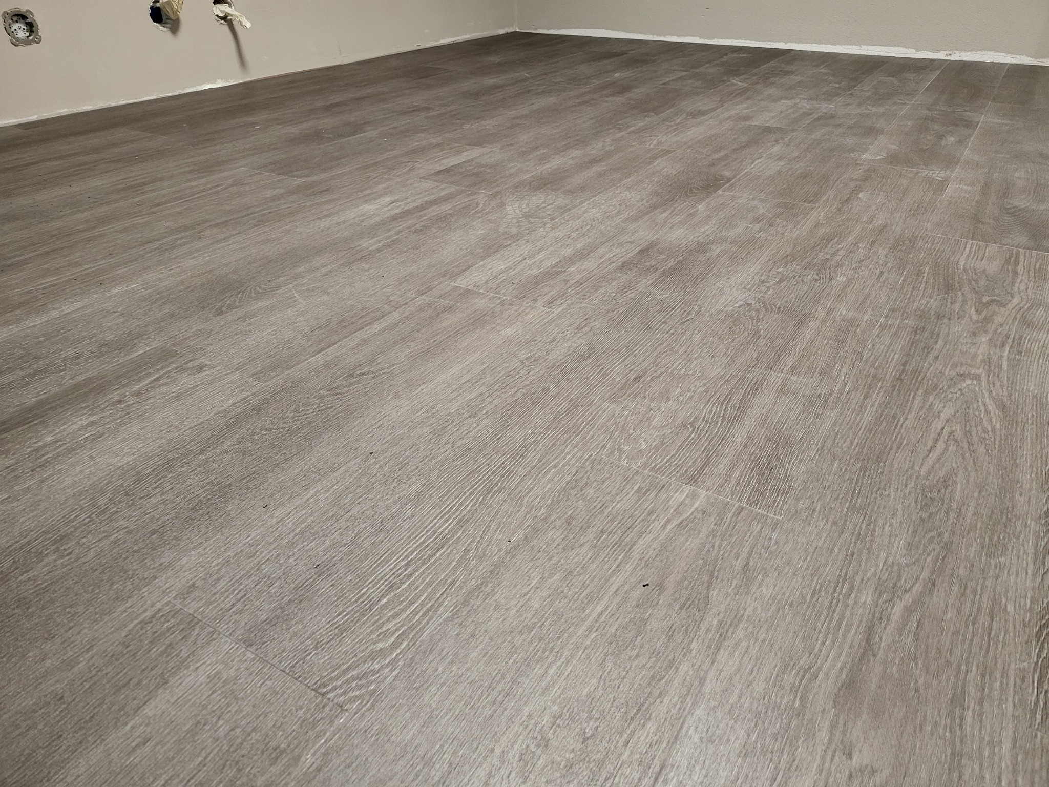 Ułożona, jasnoszara podłoga panelowa z widocznymi łączeniami, przy ścianie z wystającymi kablami elektrycznymi i gładzią na styku ściany i podłogi.