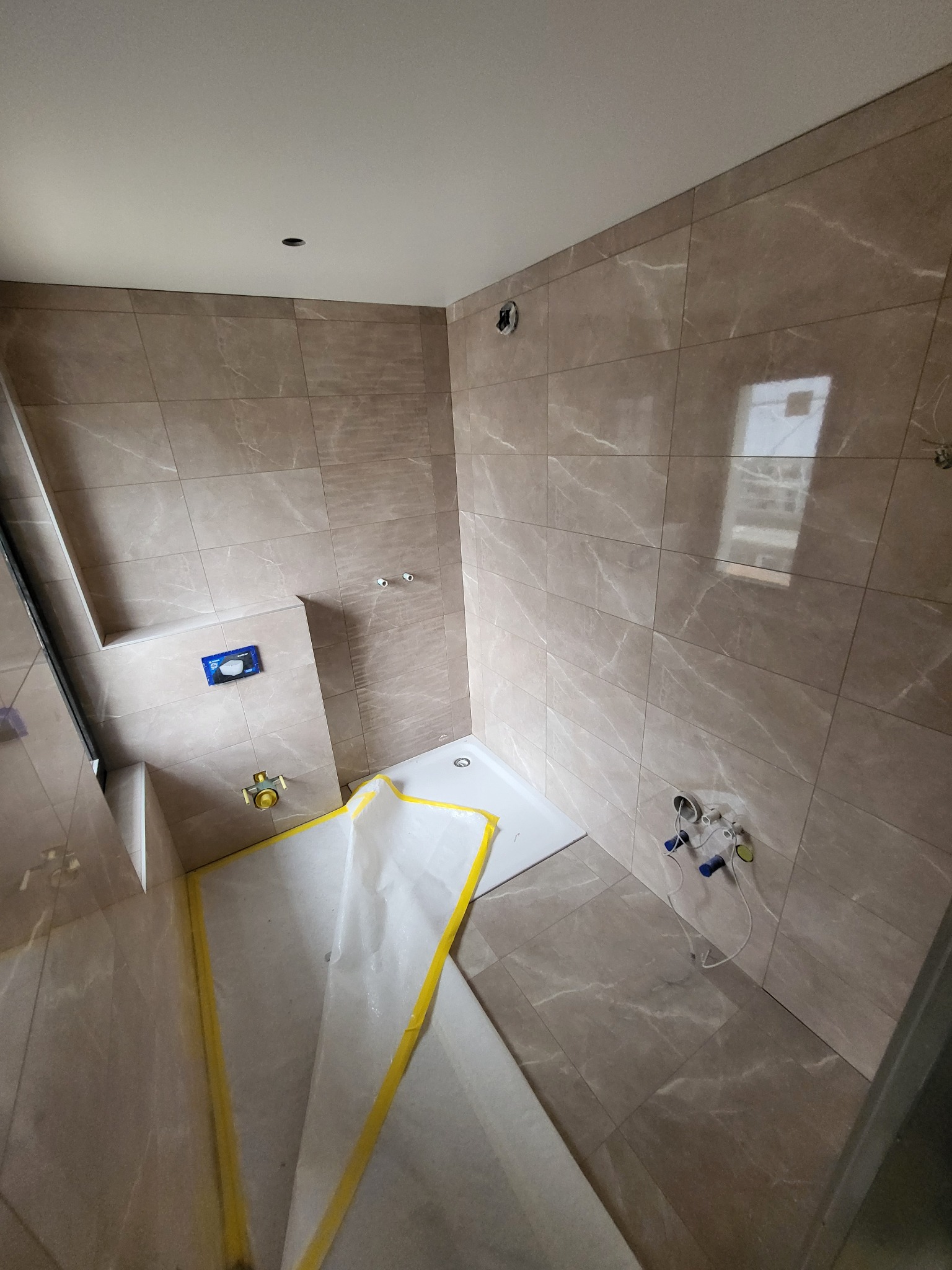 Łazienka w trakcie remontu: beżowe płytki imitujące marmur na ścianach i podłodze, biały brodzik zabezpieczony folią malarską z żółtą taśmą, widoczne instalacje wodne przygotowane do montażu armatury.