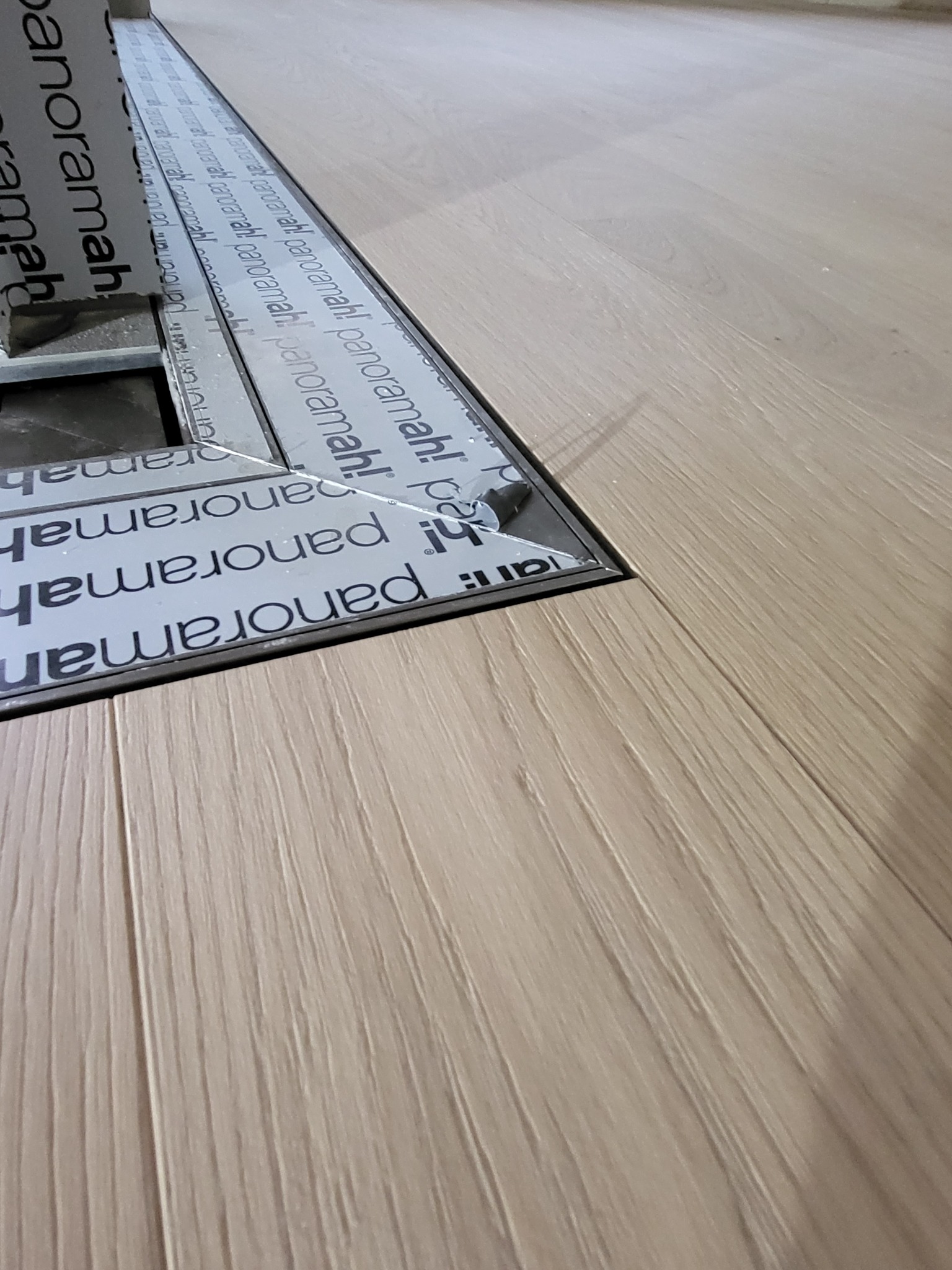 Fragment podłogi z jasnych paneli z widocznym metalowym profilem podłogowym z nadrukiem 'panoramah!', otaczającym otwór w podłodze.