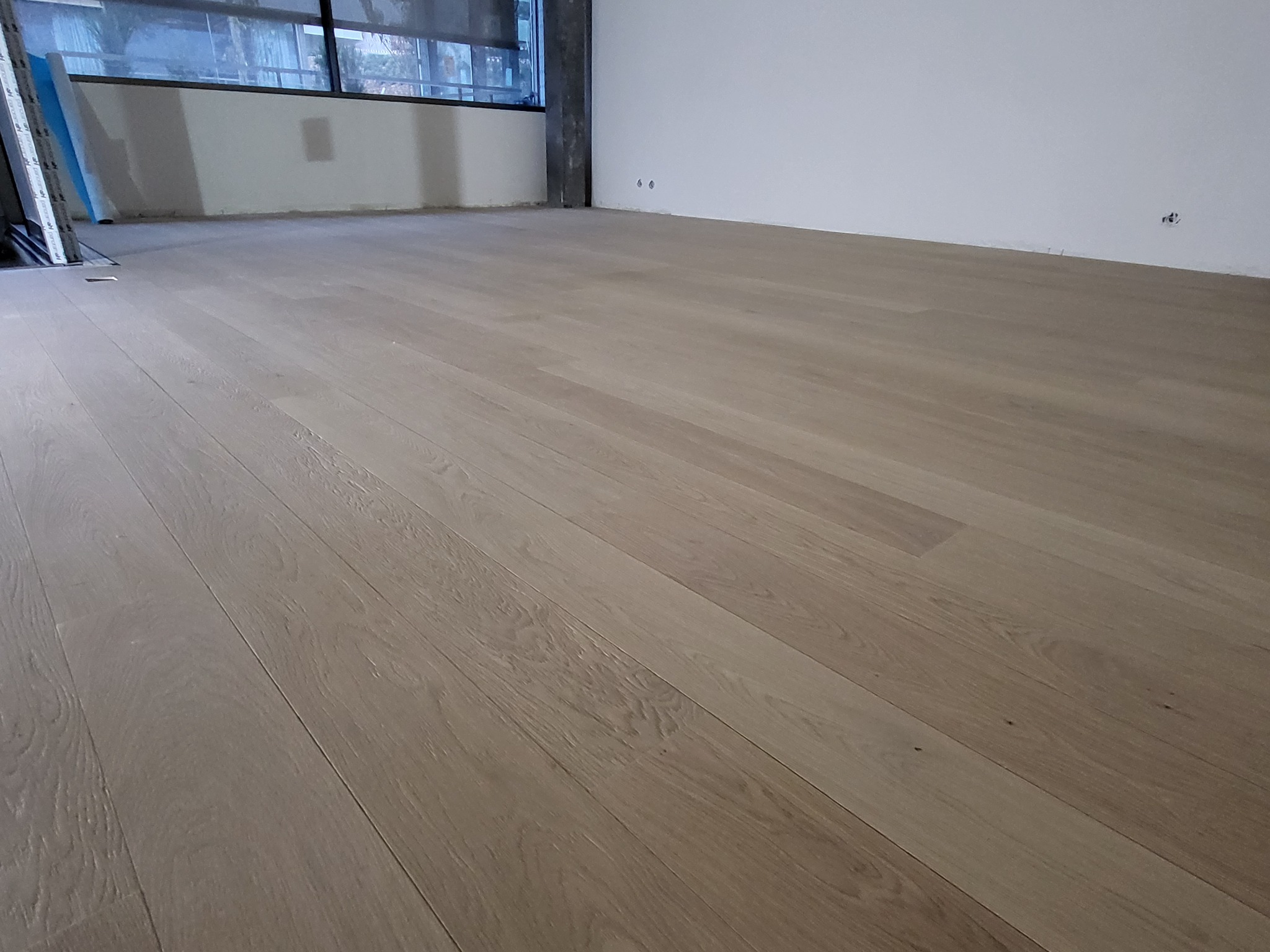 Jasna, drewniana podłoga z paneli ułożona w pustym pomieszczeniu z dużym oknem i białymi ścianami, widoczne ślady po instalacjach elektrycznych.