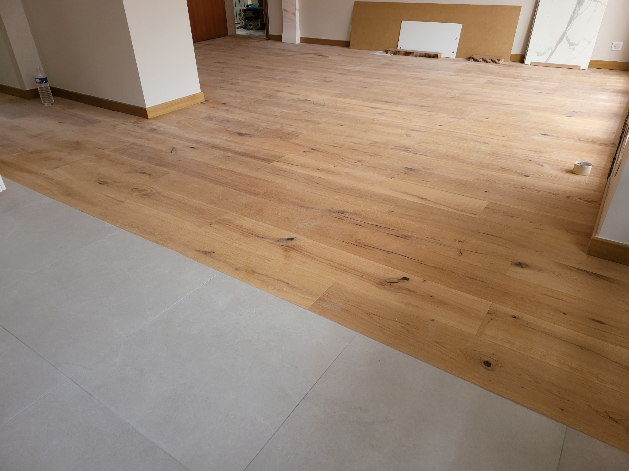 Ułożona drewniana podłoga w jasnym odcieniu, przechodząca w szare płytki podłogowe. Widoczne listwy przypodłogowe i fragment ściany. Prace wykończeniowe w Krakowie.