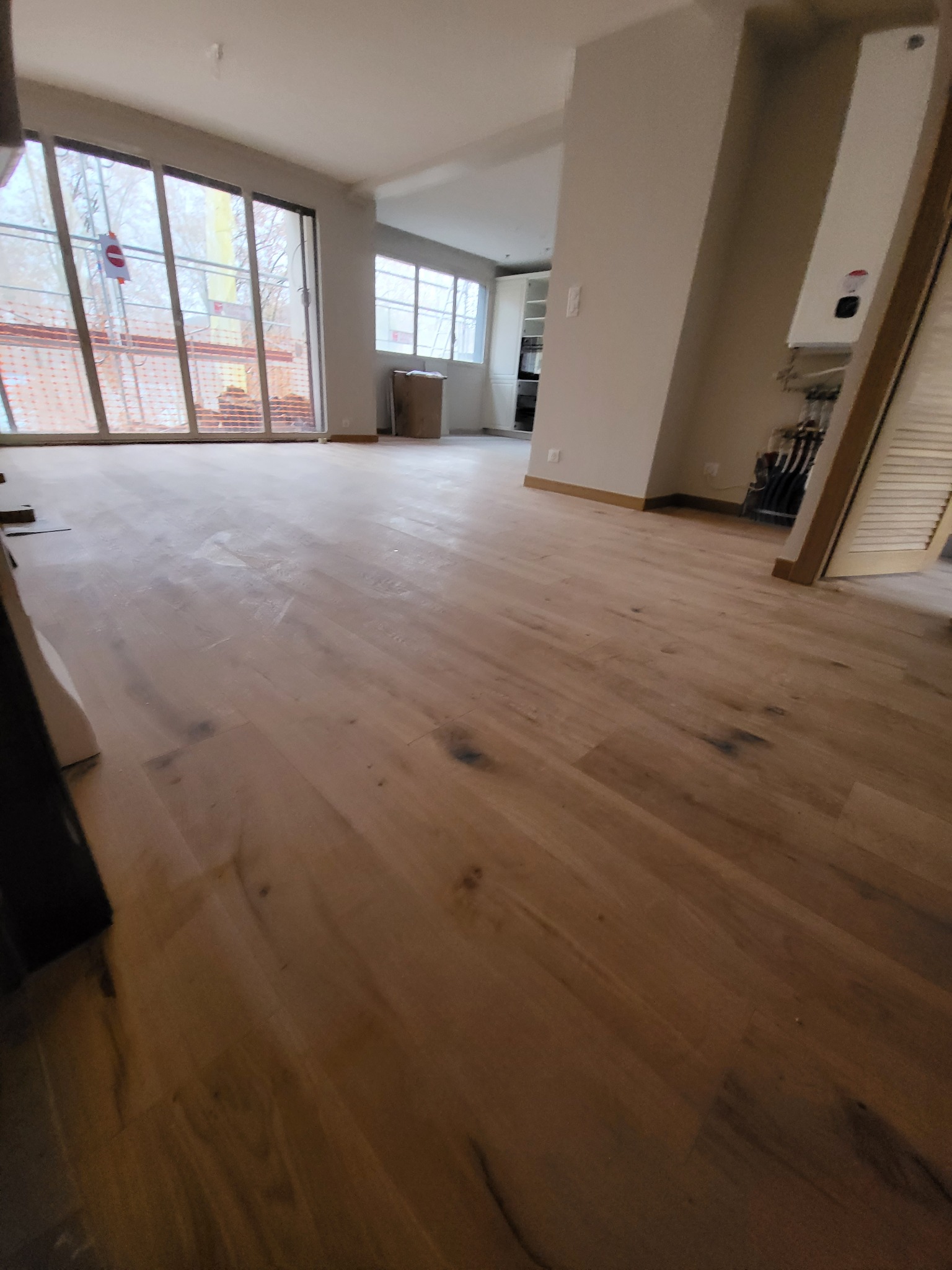 Wnętrze pokoju w trakcie prac wykończeniowych, z widoczną nową drewnianą podłogą, dużymi oknami z widokiem na rusztowania na zewnątrz oraz fragmentem instalacji grzewczej na ścianie.