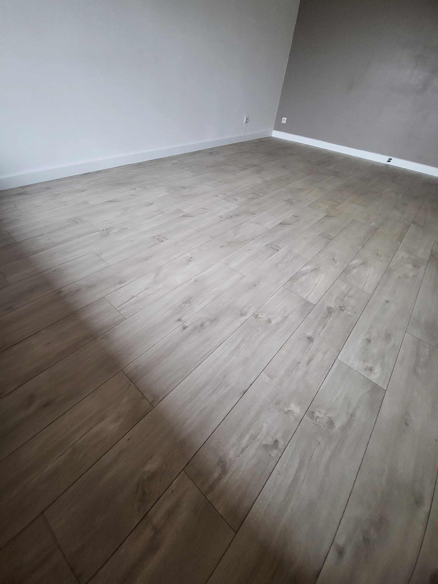 Pusty pokój po remoncie z nową podłogą z paneli w odcieniach szarości, ściany pomalowane na biało i szaro, widoczne gniazdka elektryczne.